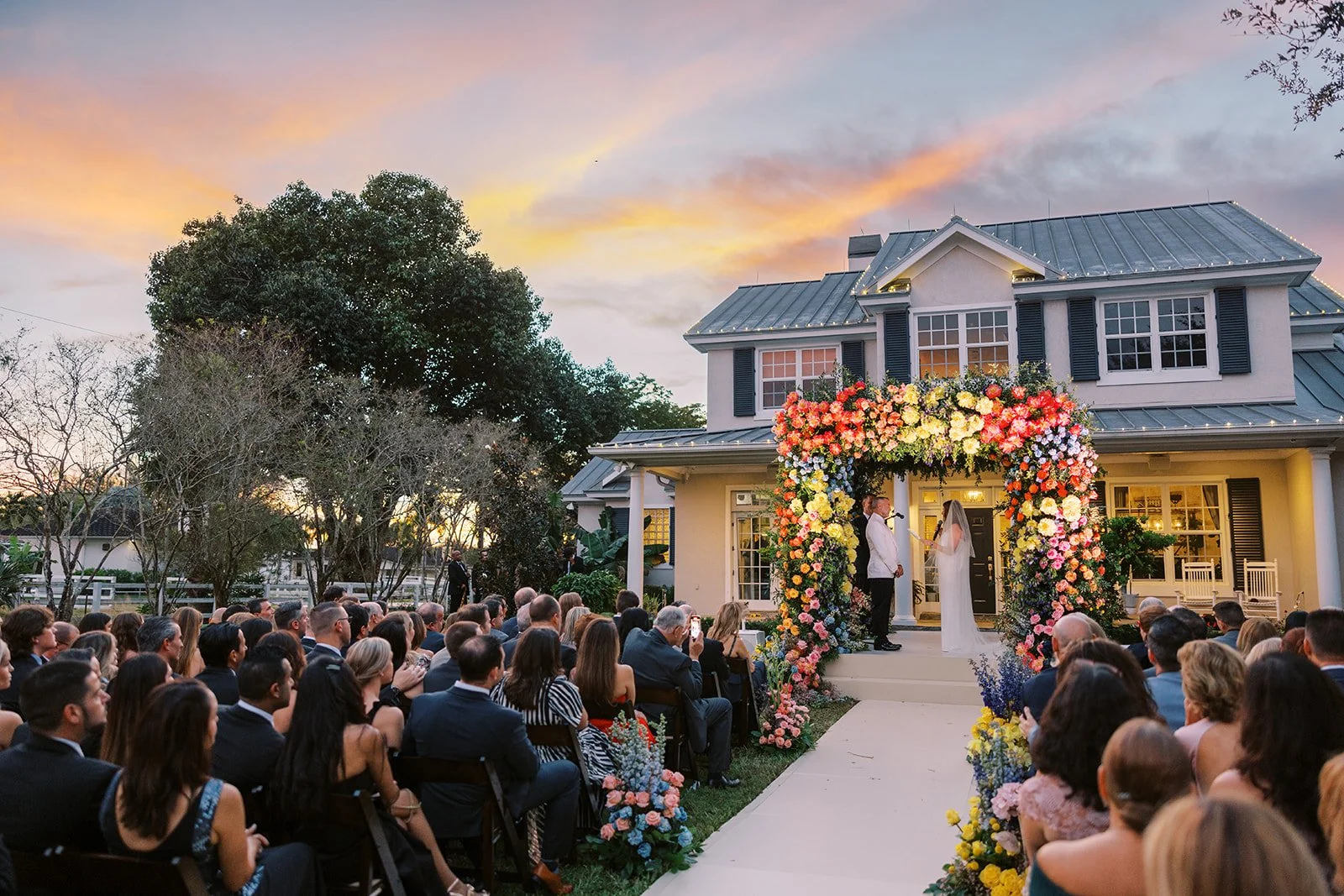 A wedding ceremony taking place outdoors in front of a large house decorated with a floral arch. The bride and groom are standing under the arch, exchanging vows, with guests seated facing them during sunset.