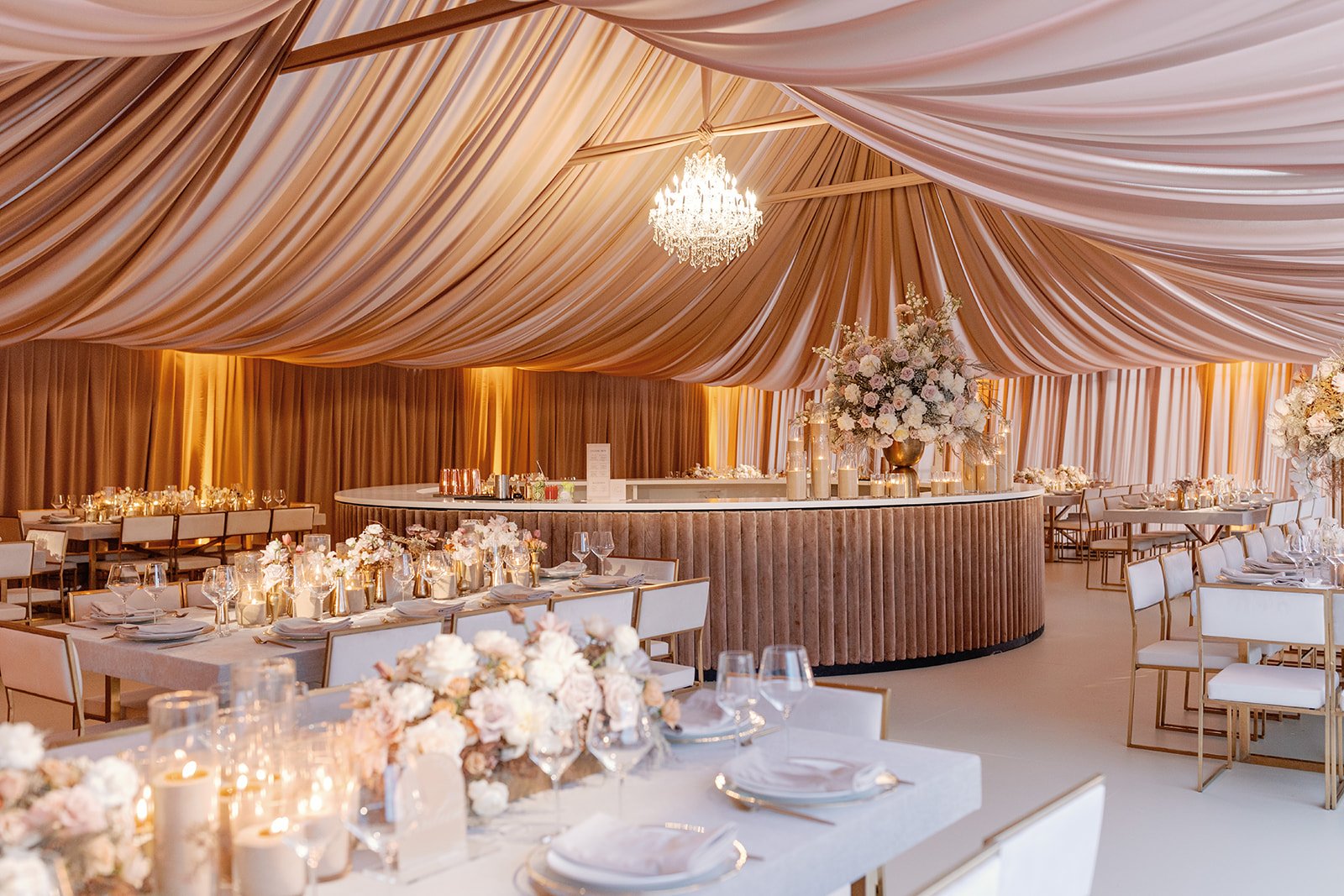 Elegant event space decorated with floral centerpieces, candles, and warm lighting, featuring draped ceiling fabric and a chandelier.