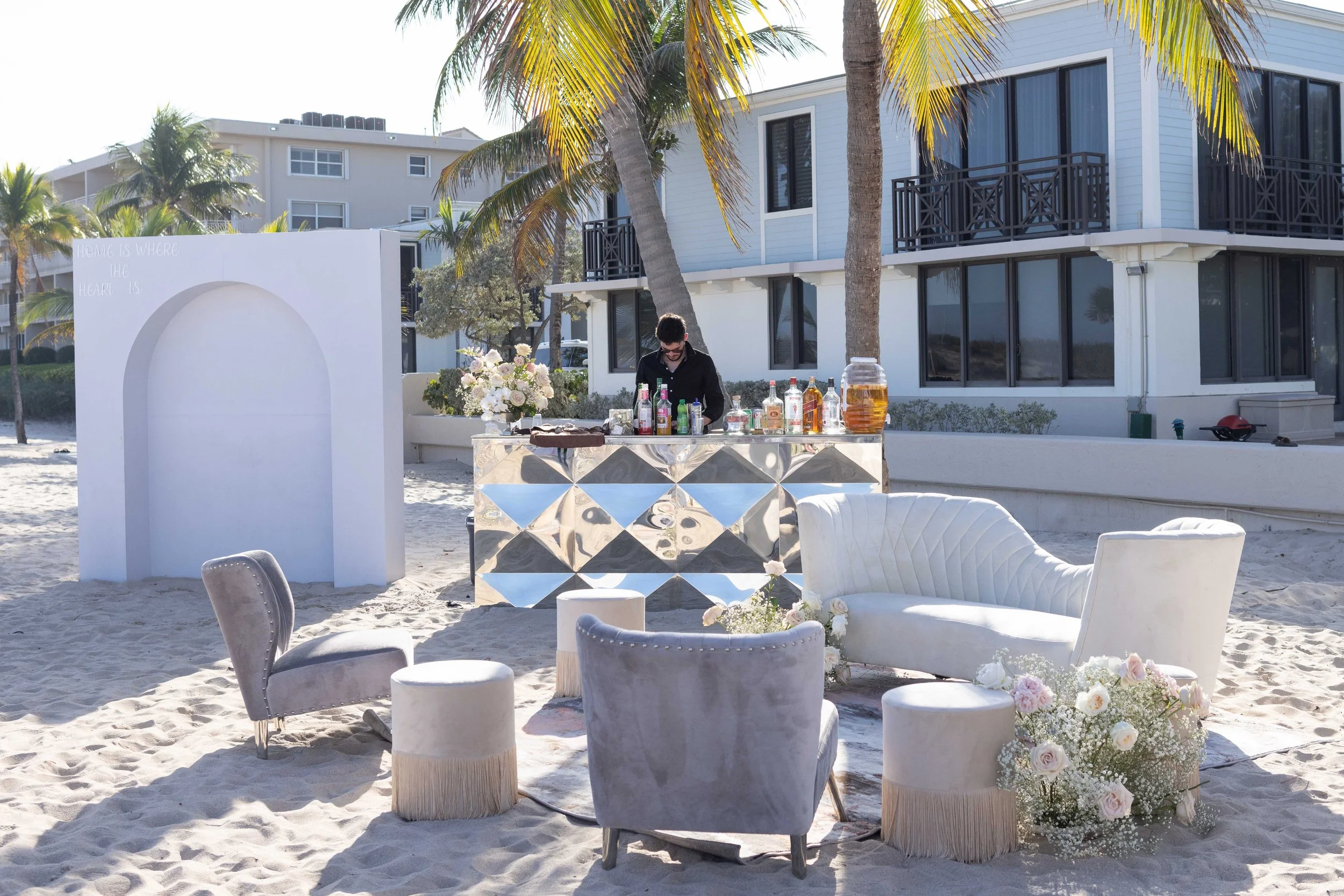A beach setup with white chairs and a drink station featuring various bottles of alcohol and a large flower arrangement, surrounded by palm trees and a modern building in the background.