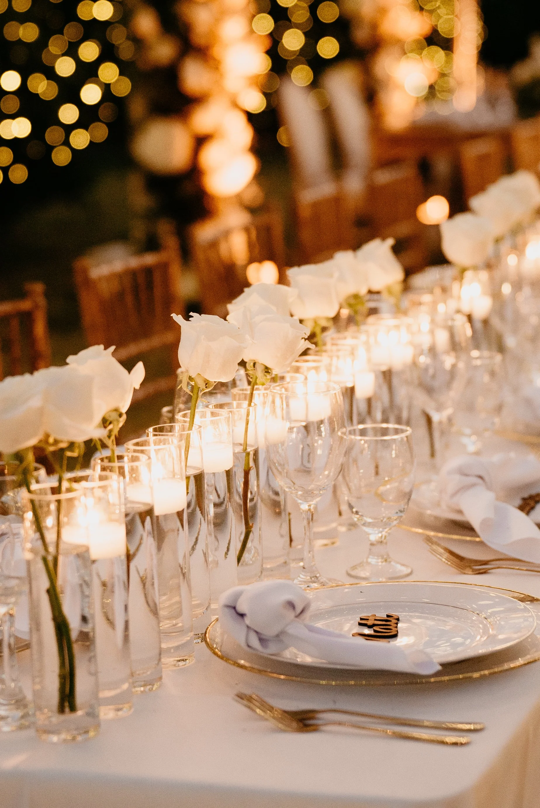 A decorated dinner table with white flowers in glass vases, lit candles, and elegant place settings, including plates, napkins, and utensils, under string lights in a warm, festive ambiance.