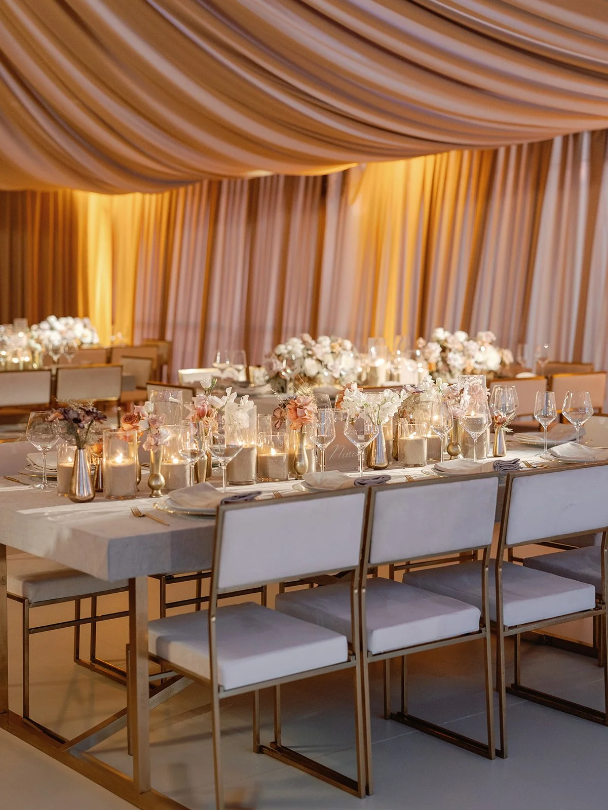 Elegant banquet table decorated with candles, flowers, and glassware under draped, warm-colored curtains.