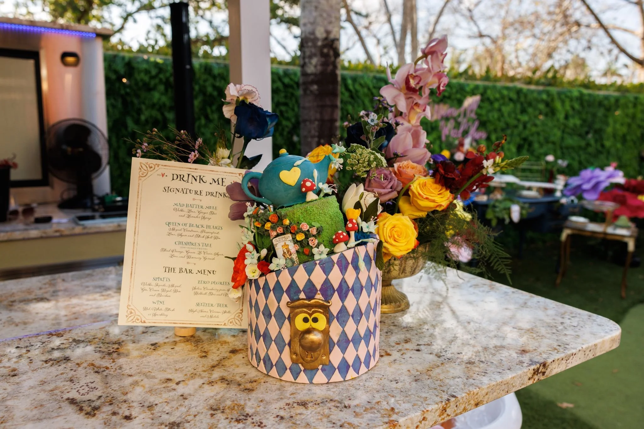 Colorful table centerpiece with an assortment of flowers including roses and orchids, a blue teapot cake decorated with flowers and mushrooms, and a menu card, set outdoors with trees and a hedge in the background.