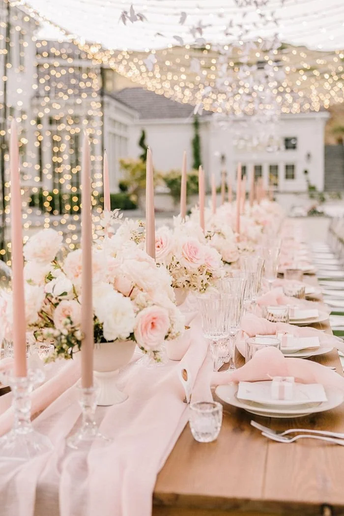Elegant outdoor wedding table decorated with pink and white flowers, pink candles, pink napkins, and glassware under string lights.