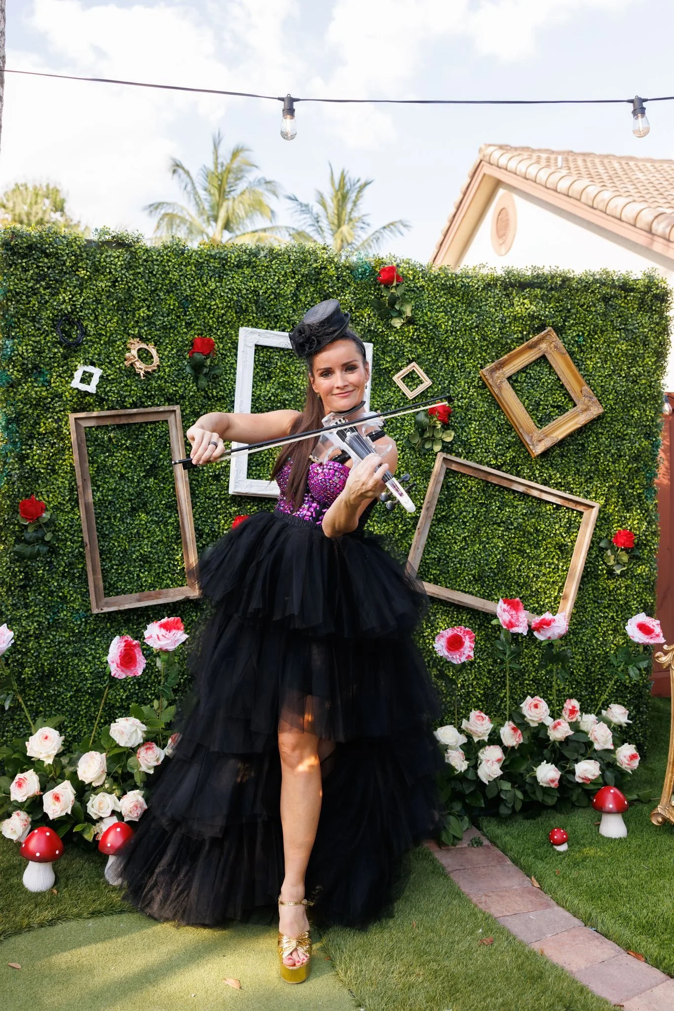 A woman in a black dress and gold platform shoes holding a violin, standing in front of a decorated green hedge with pink and white flowers, framed pictures, and mushroom-shaped lawn ornaments.