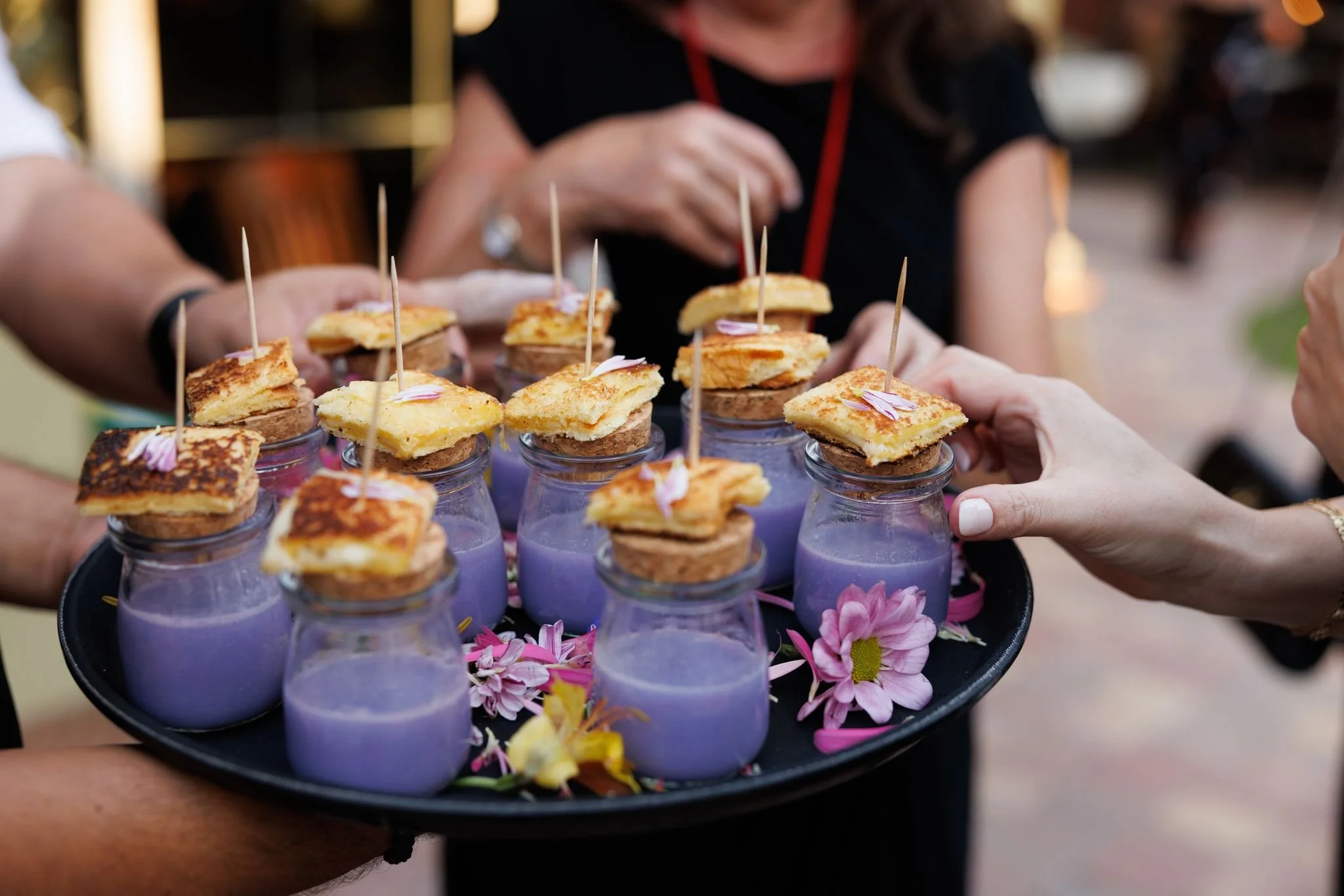 A tray of small glasses filled with purple beverages, each topped with pieces of flatbread or toast and pink flower petals, being handed around at an outdoor social gathering amid some floral decorations.