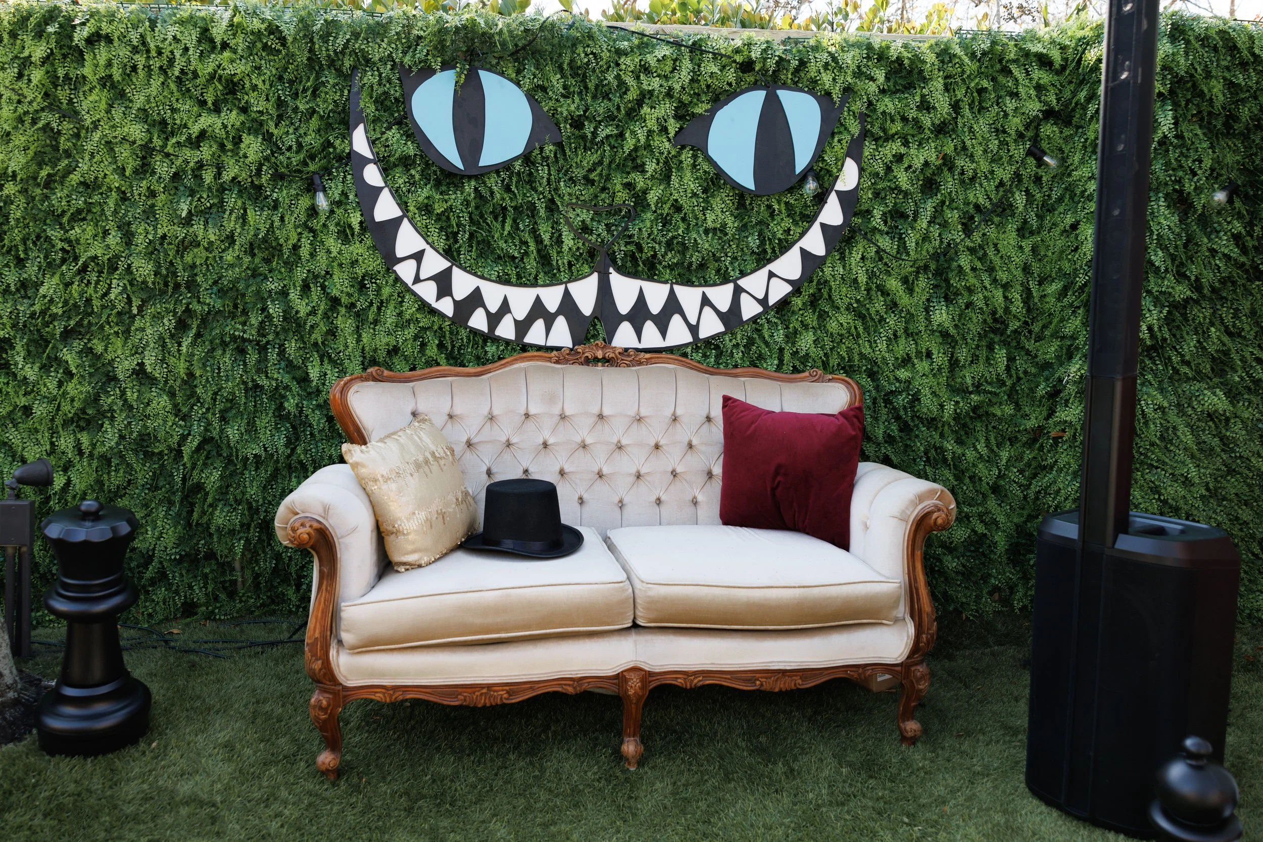Vintage cream-colored sofa with wooden trim, two pillows (one silver, one red), a black top hat, and a black small table in front, set outdoors in front of a green hedge wall with a smiling face design, string lights, and greenery above.