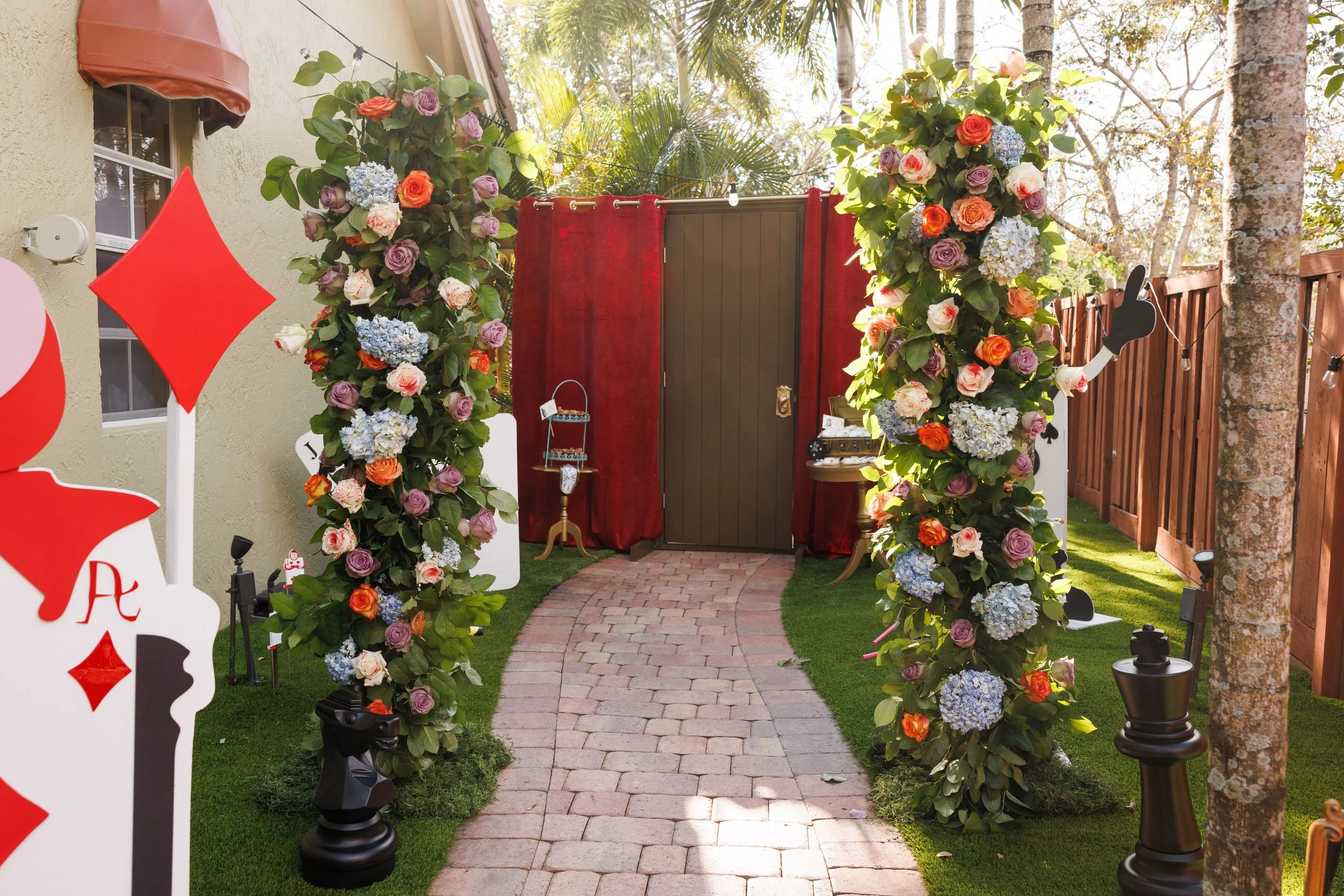 Decorative archway of colorful roses, hydrangeas, and greenery over a brick pathway leading to a door, with outdoor string lights and trees in background.