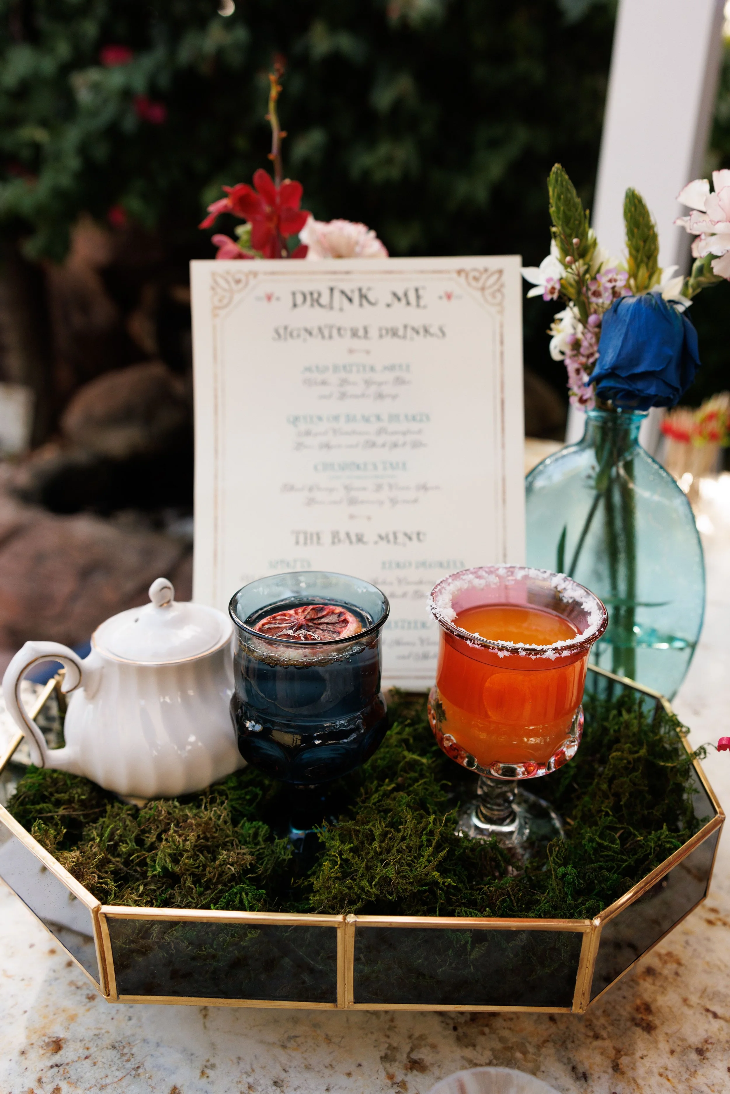 A decorative tray with colorful drinks, a teapot, and a sign that reads 'Drink Me' surrounded by flowers and greenery.