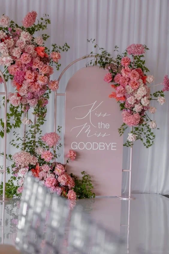 A wedding or farewell event setup with a pink sign that reads 'Kiss the Miss Goodbye' surrounded by pink and white floral arrangements on a white background.