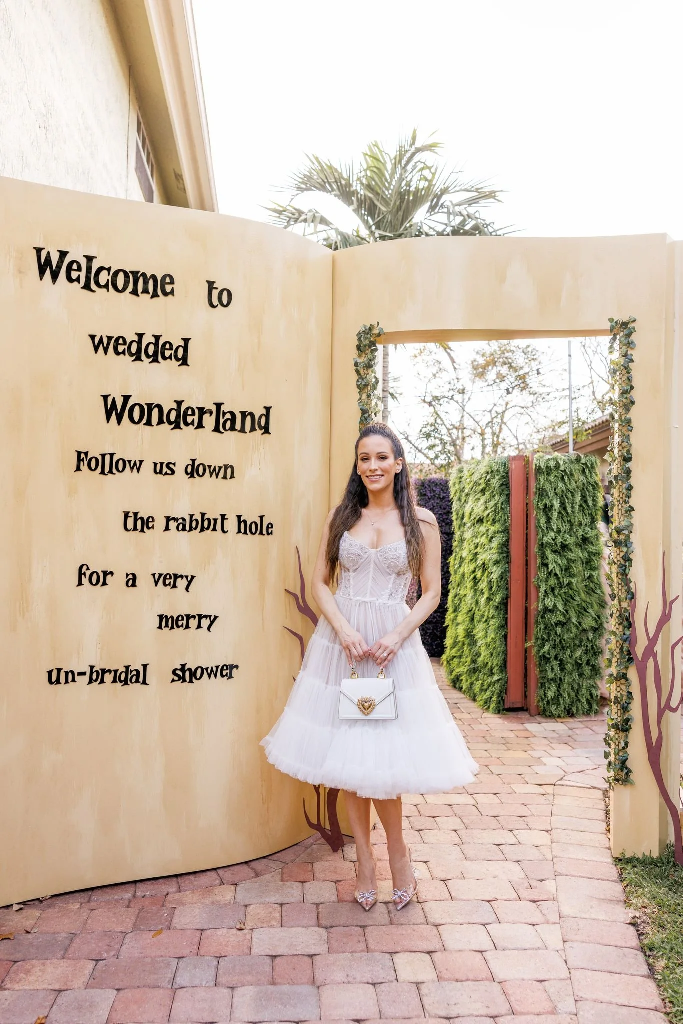 A smiling bride in a wedding dress standing on a brick pathway outside at a wedding venue. She holds a white handbag with a gold heart embellishment. A whimsical, large, open book-shaped sign with black text welcomes guests to a wedding and mentions 