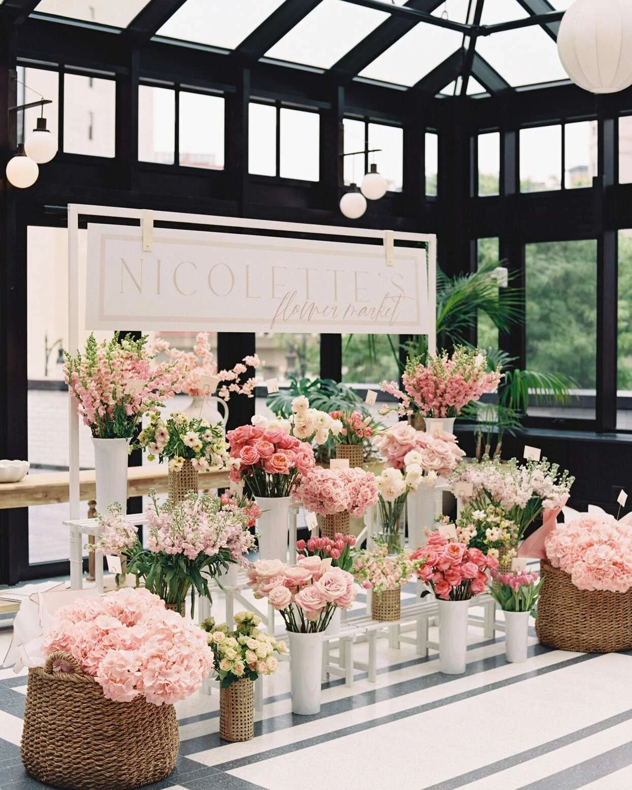Display of pink and white flowers in vases at a floral shop named Nicoletti Flower Market, inside a glass-enclosed building with black framing and hanging globe lights.