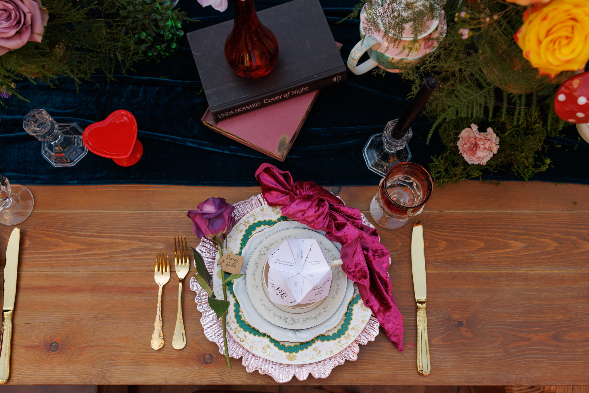 A decorated table with fine china, gold cutlery, a pink velvet napkin, and a flower. There are various flowers and greenery in the background, along with a small white takeout box with a fork and a label reading "Audrey Saltz" on the plate. Gold and 