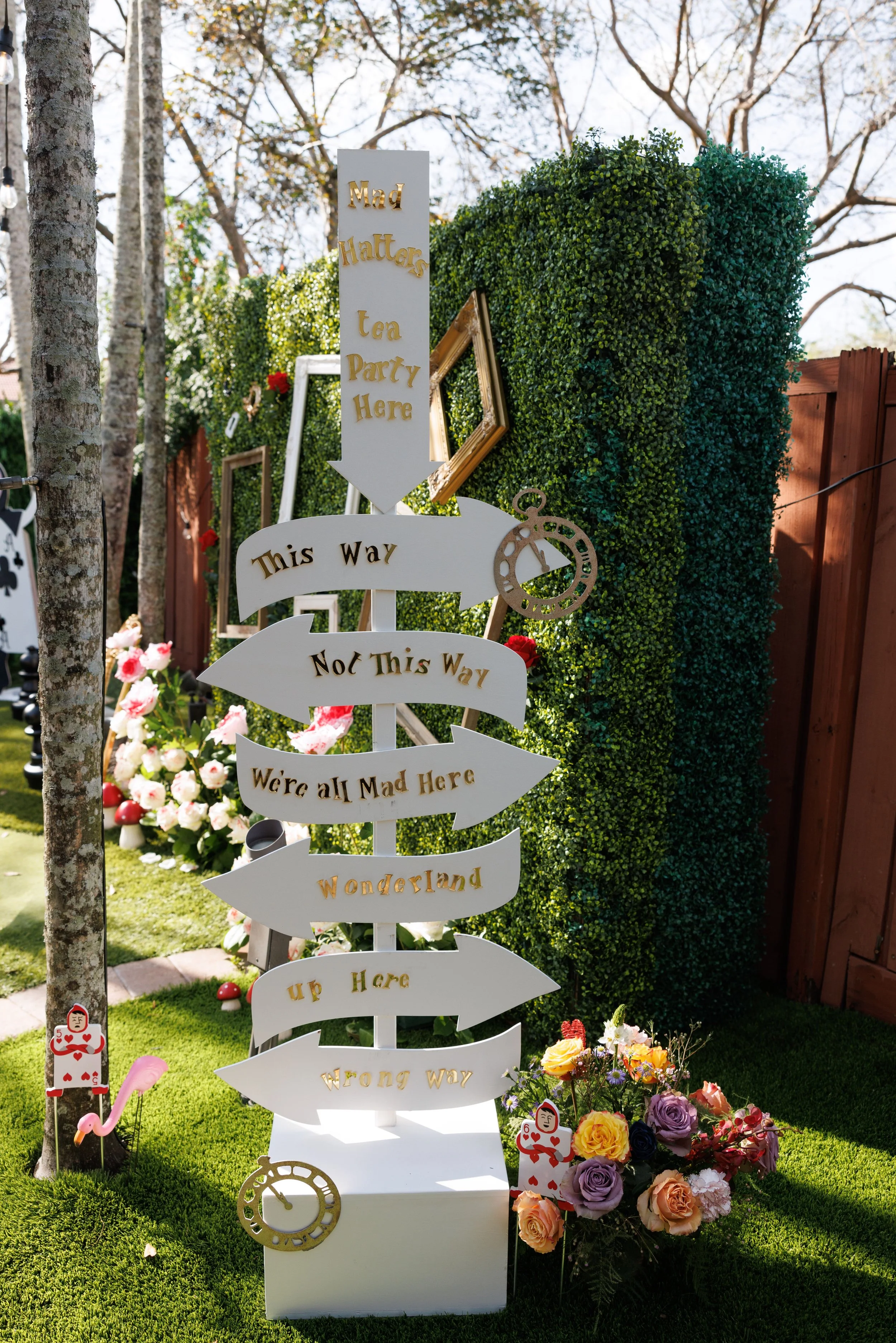 Signpost with arrows pointing in different directions, each labeled with whimsical text such as "Mad Hatters Tea Party Here," "This Way," "Not This Way," "We're all Mad Here," "Wong Way," and "up Here." The signpost is positioned outdoors in a garden