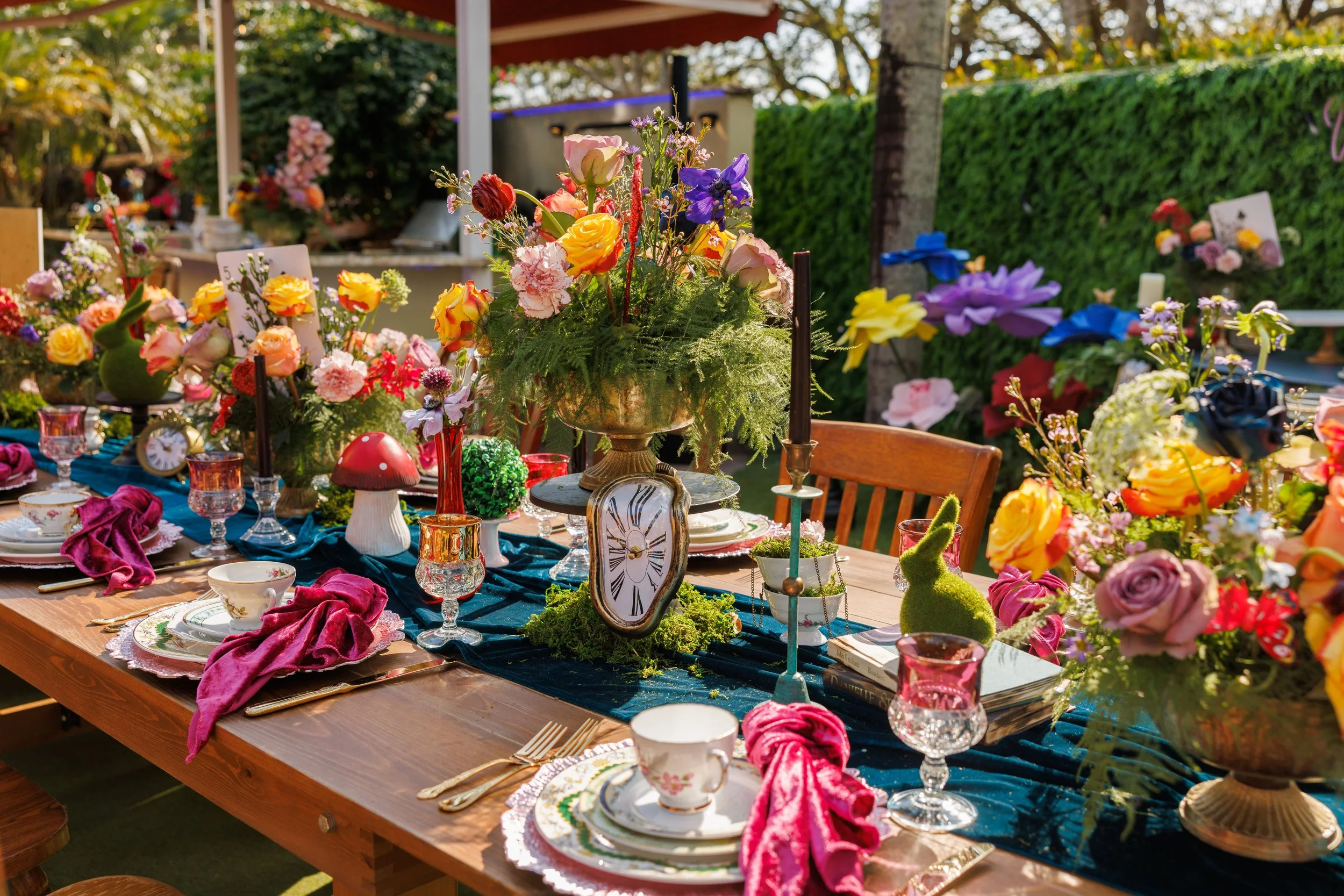 A long wooden outdoor table set for a celebration with various floral arrangements, colorful glassware, and decorative items, surrounded by trees and greenery.