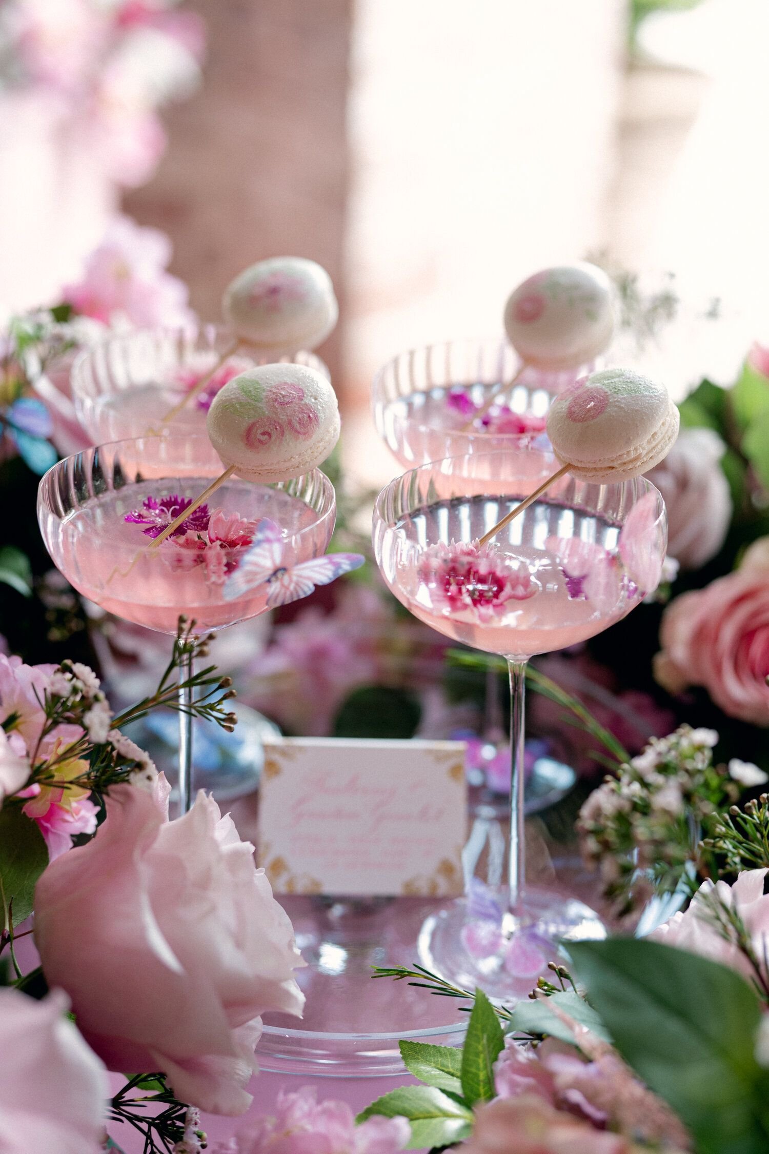 Close-up of pink macaron desserts with floral decoration in glass cups, surrounded by pink and white flowers with greenery.