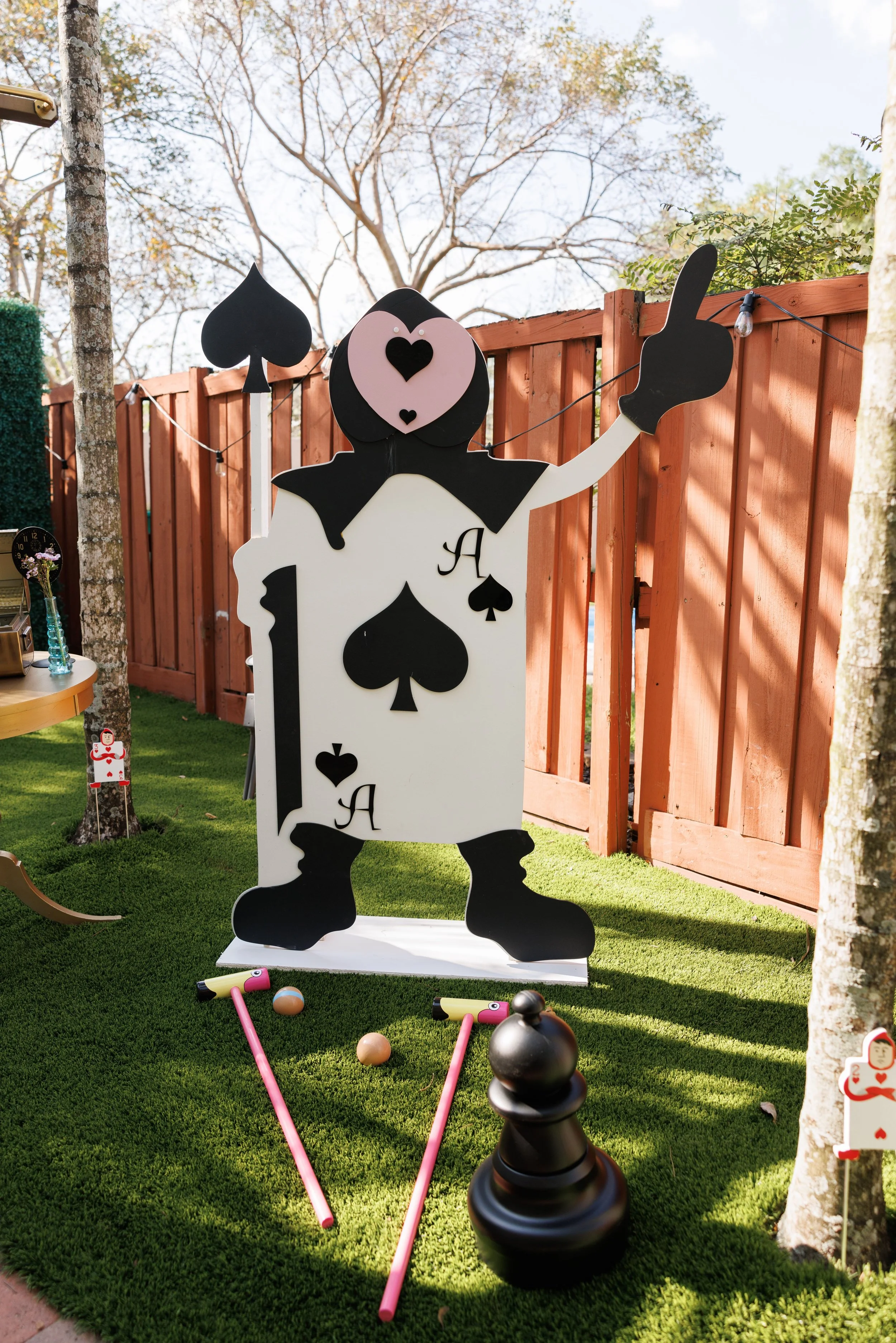Outdoor yard scene with large decorative cutout of a cartoonish dog made from big playing card shapes, holding up a peace sign with its paw. There are small toys including pink golf clubs, golf balls, and a black bowling ball on green artificial gras