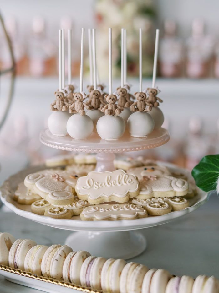 Cake pops decorated with teddy bear toppers, cookies with the name 'Isabelle' written on them, and macarons on a white serving stand for a celebration.