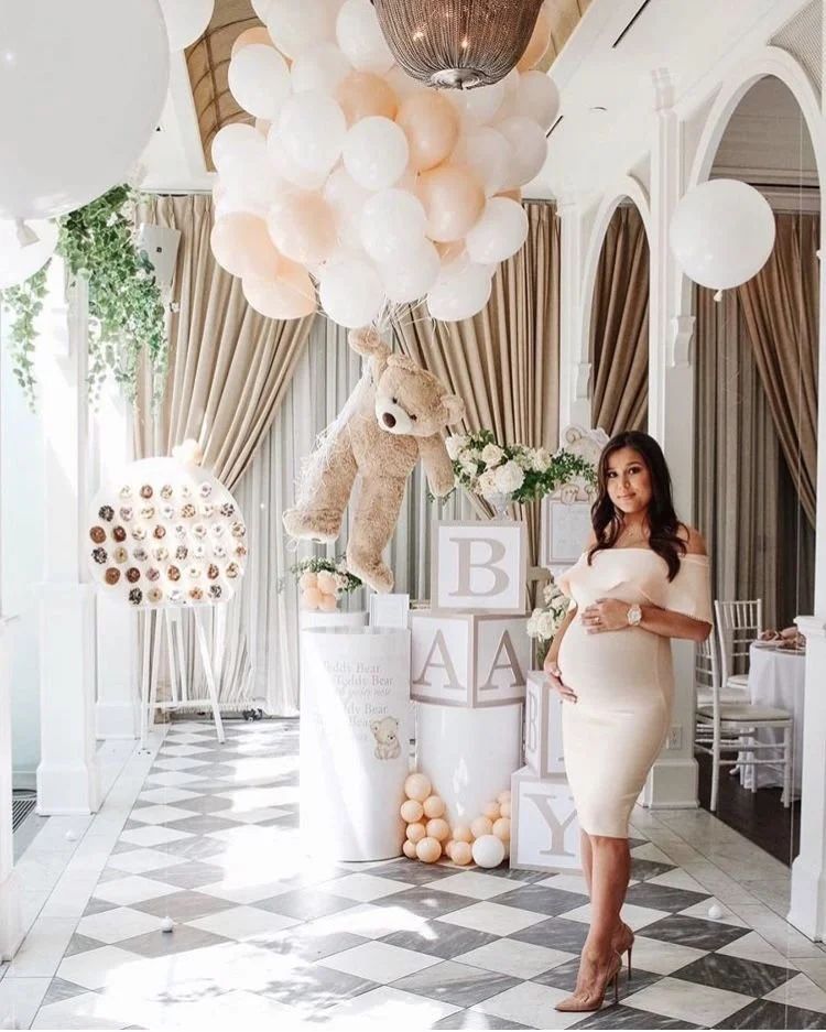 A woman in a cream-colored off-shoulder dress standing in a decorated room with balloons, teddy bear, and pastel-colored blocks spelling 'BABY' for a gender reveal or baby shower celebration.