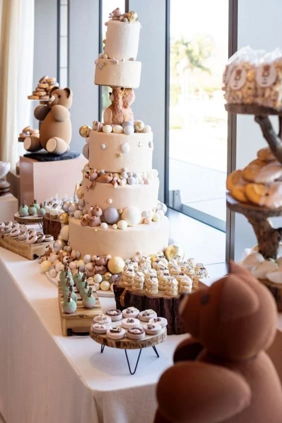 A multi-tiered cake decorated with pastel-colored ornaments and surrounded by various cupcakes and desserts on a white table, with teddy bear decorations nearby.