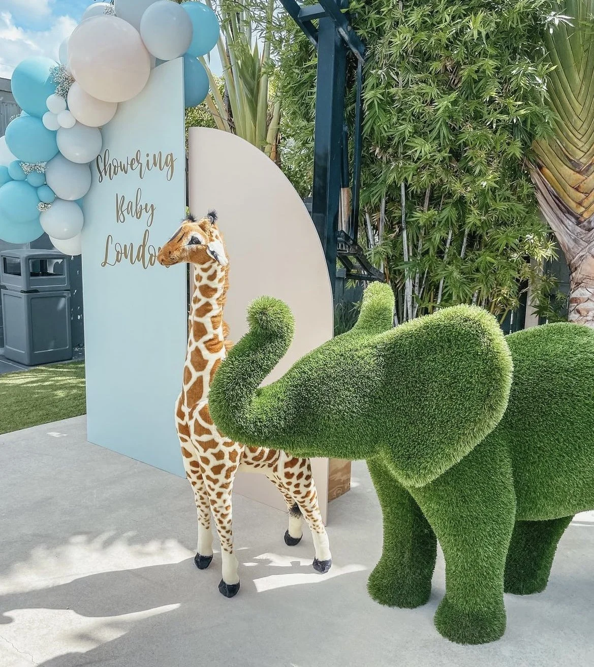 Decorative setup for a baby shower with a giraffe plush toy, a green elephant-shaped topiary, and a sign that reads "Showering Baby London" with balloons in light blue, white, and gray, outdoors with greenery in the background.