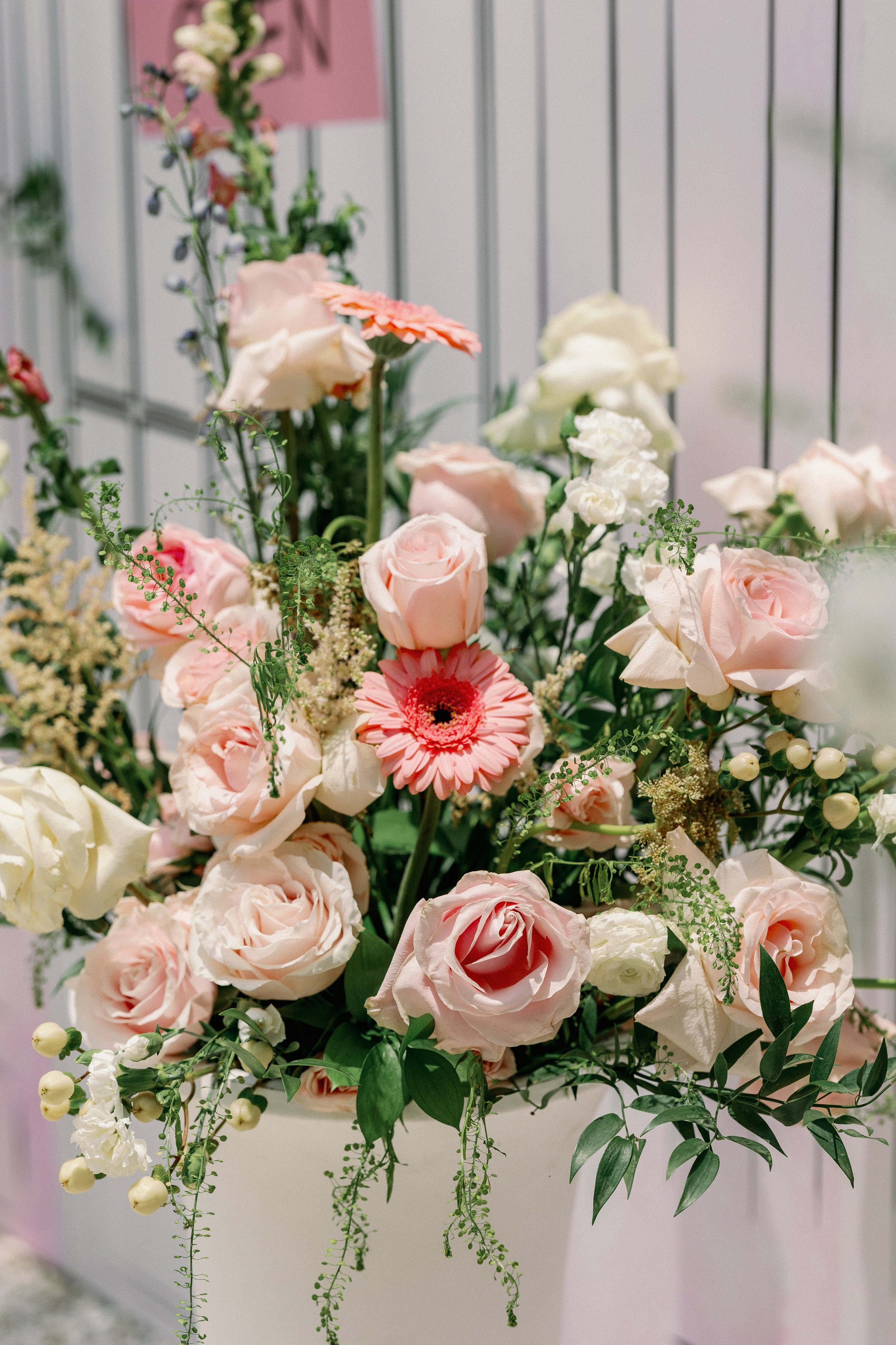A bouquet of pink and white roses, white and pink gerbera daisies, and various green foliage arranged in front of a pink and white background.