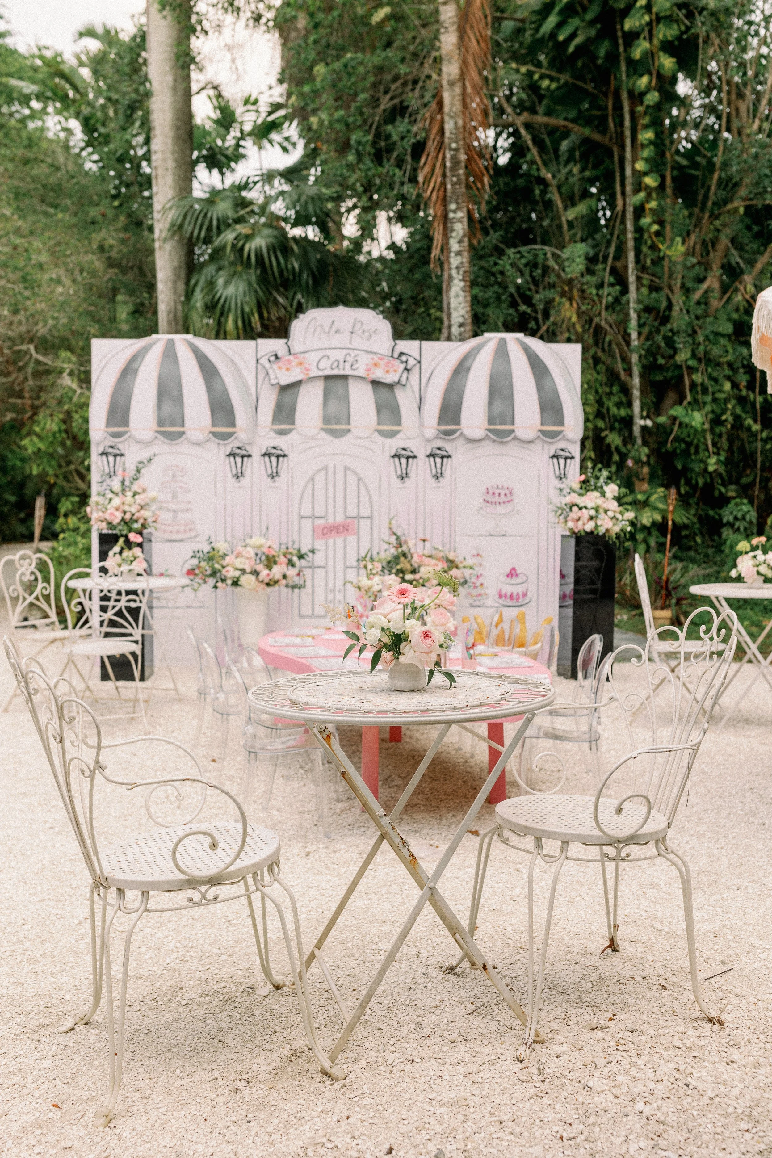 A decorated outdoor cafe area with white wrought iron chairs and tables, pink and white floral arrangements, and a backdrop with a pastel pink and white striped awning and a sign that reads 'Make Rose Café.' There are trees and greenery in the backgr
