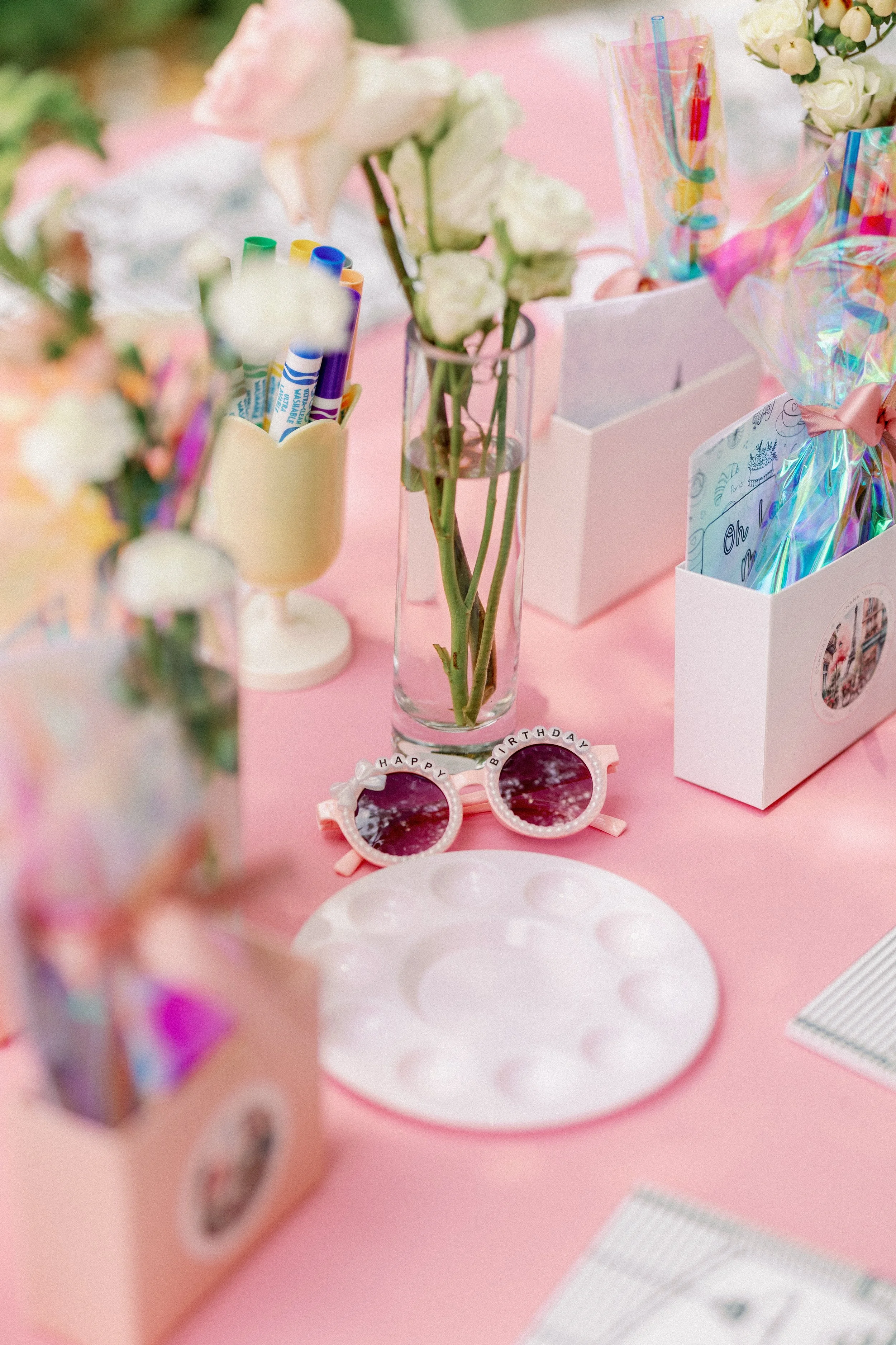 Birthday party table decorated with pink and white flowers, rainbow-colored straws, pink sunglasses with 'Happy Birthday' written on the frames, and gift boxes wrapped in holographic paper on a pink surface.