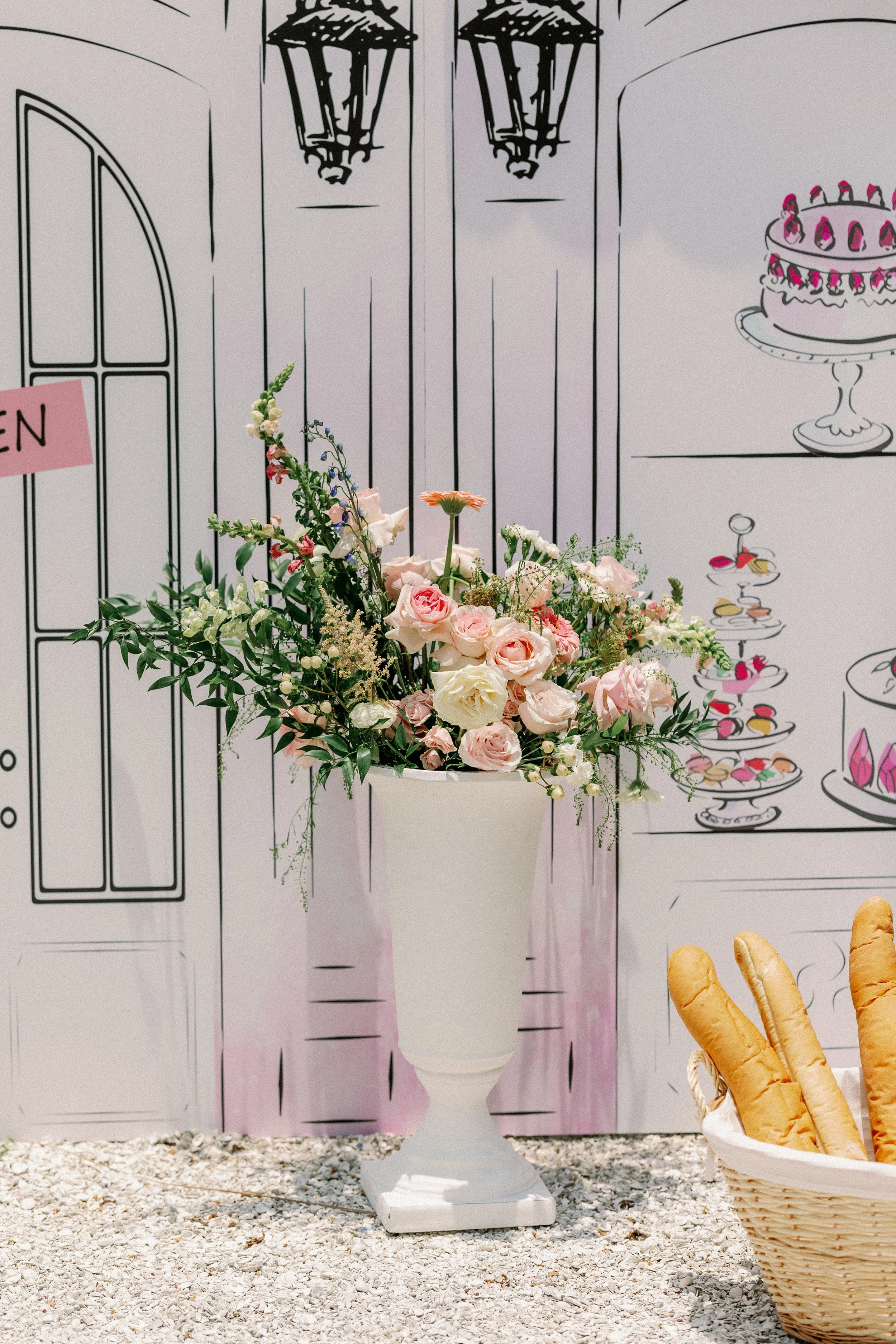 A large white pedestal holding a bouquet of pink, white, and peach flowers with greenery, positioned in front of a black-and-white illustrated backdrop of a door, windows, and a cake on a stand. To the right, a basket of baguettes is visible.