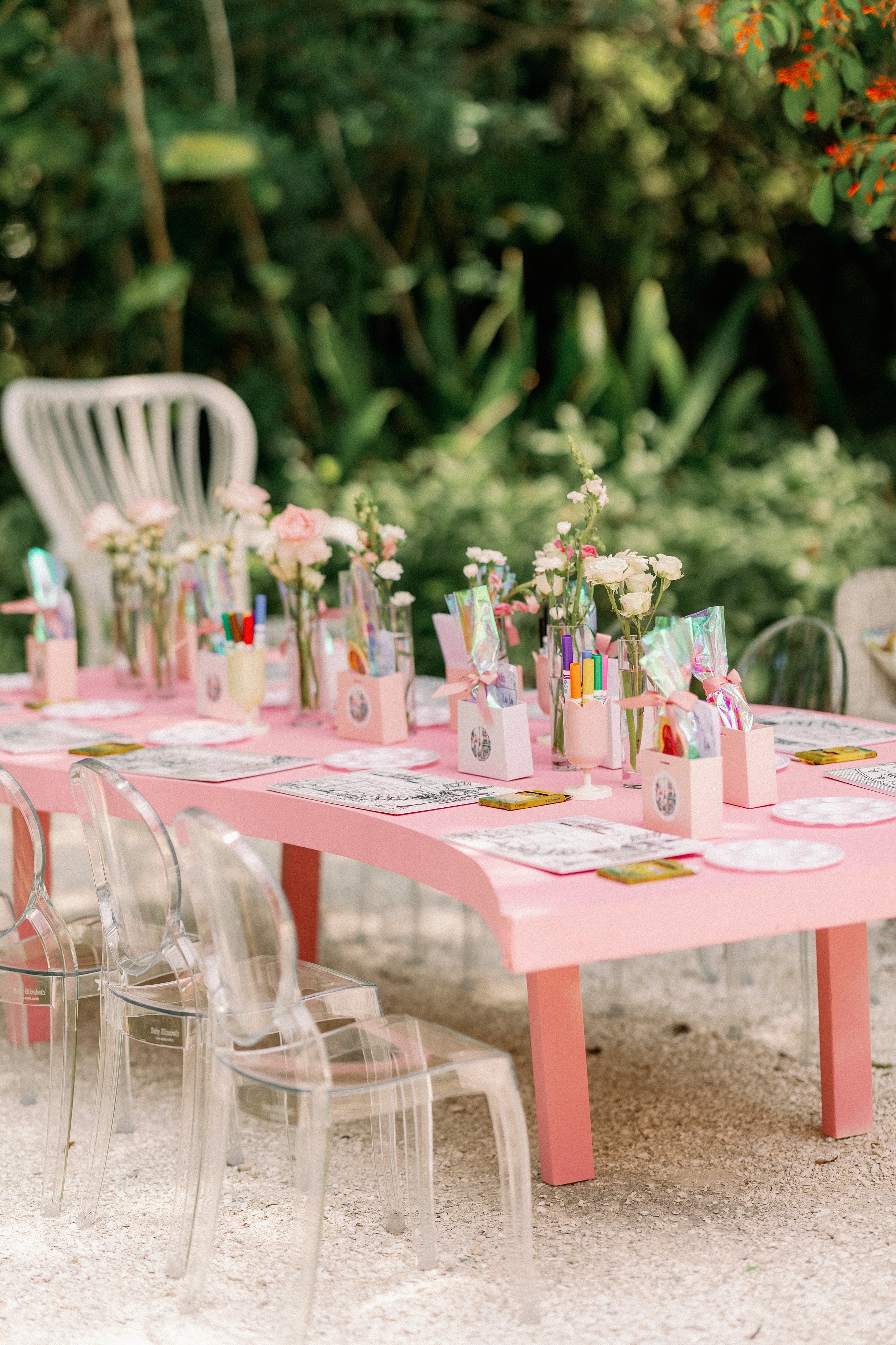A pink outdoor table decorated for a celebration, with glass jars filled with flowers, surrounded by clear acrylic chairs, set on gravel with greenery in the background.