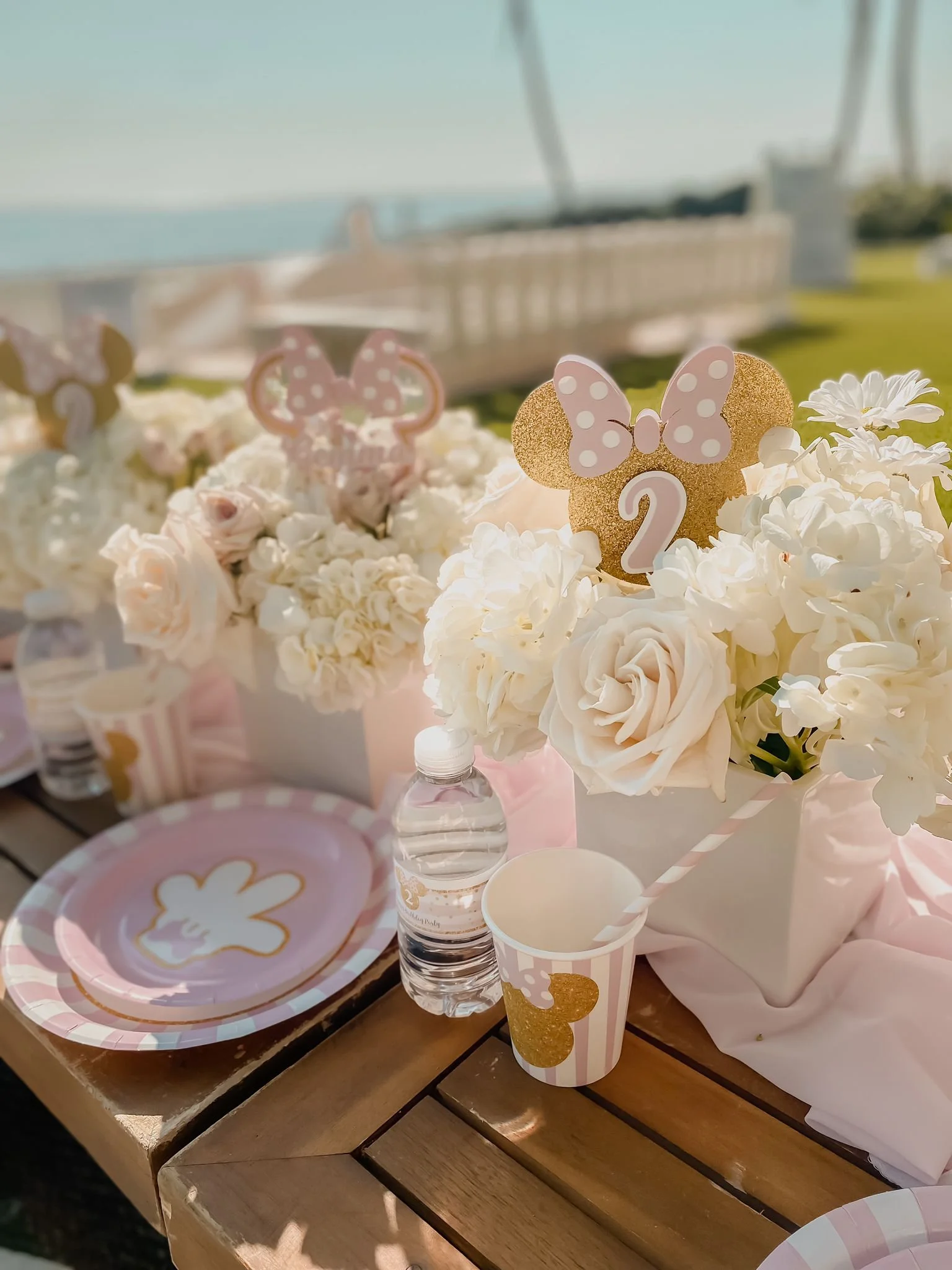 Decorations for a celebration, including flower arrangements, paper plates and cups with pink and gold butterfly and polka dot motifs, a small water bottle, and a card on a wooden table, with a blurred outdoor background.