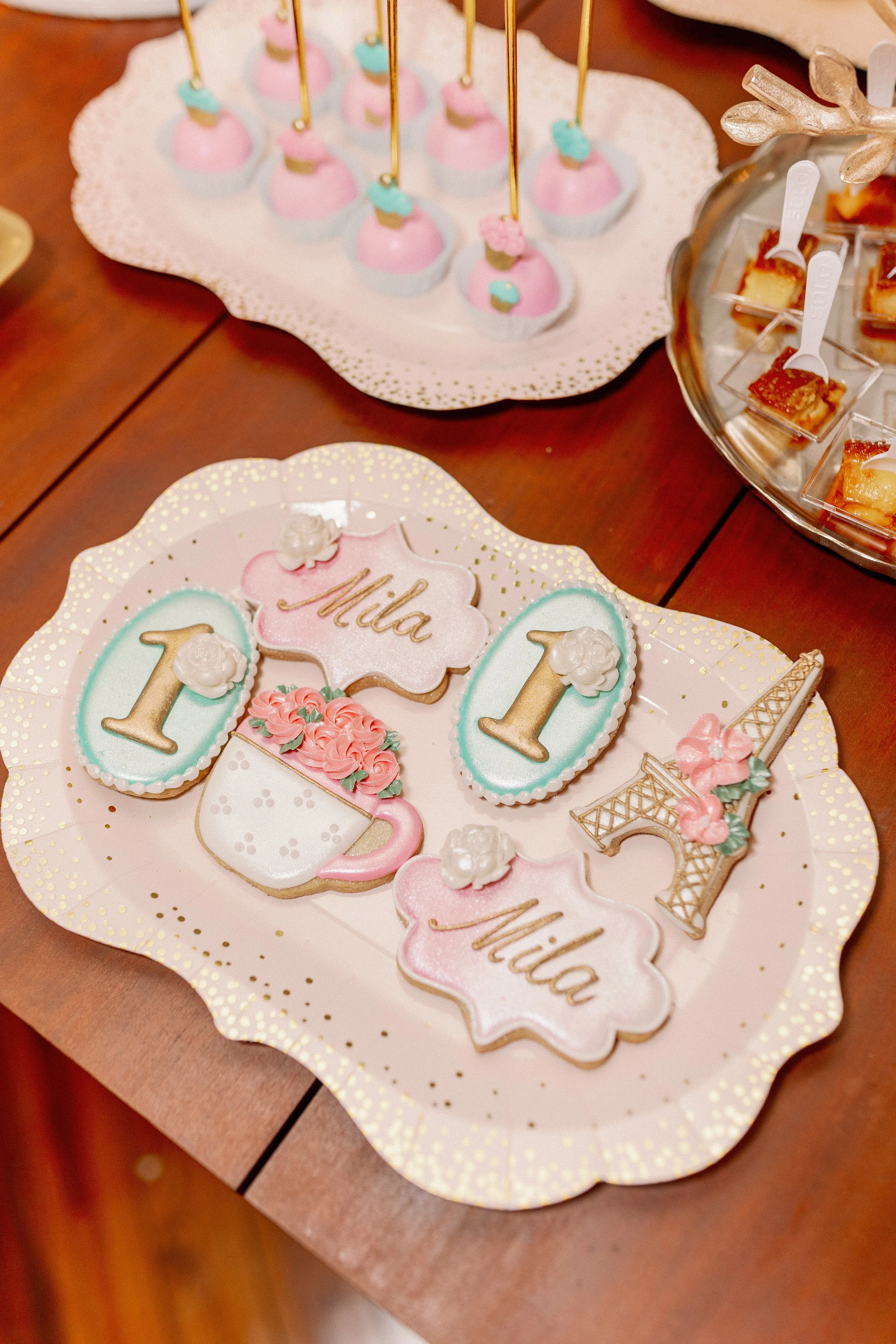 Decorative cookies on a party tray with pastel colors. The cookies spell out 'Mila' and '1' indicating a first birthday celebration, with themed shapes including champagne glasses, a Eiffel Tower, and flowers.