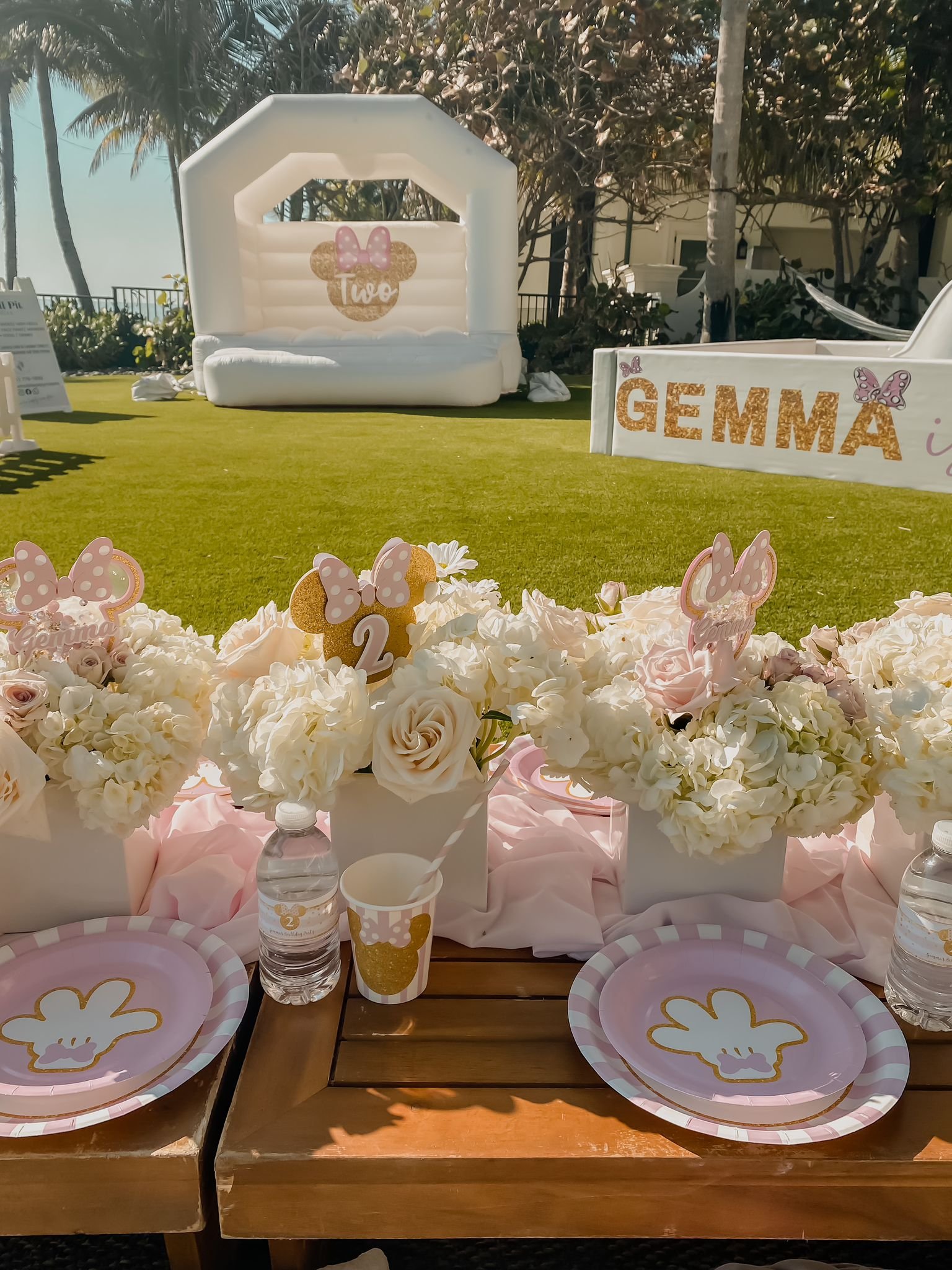Decorative table setup for a birthday celebration, featuring floral arrangements with white and pink flowers, personalized plates with bunny illustrations, cups with a small bunny logo, and water bottles. In the background, there is a white inflatabl