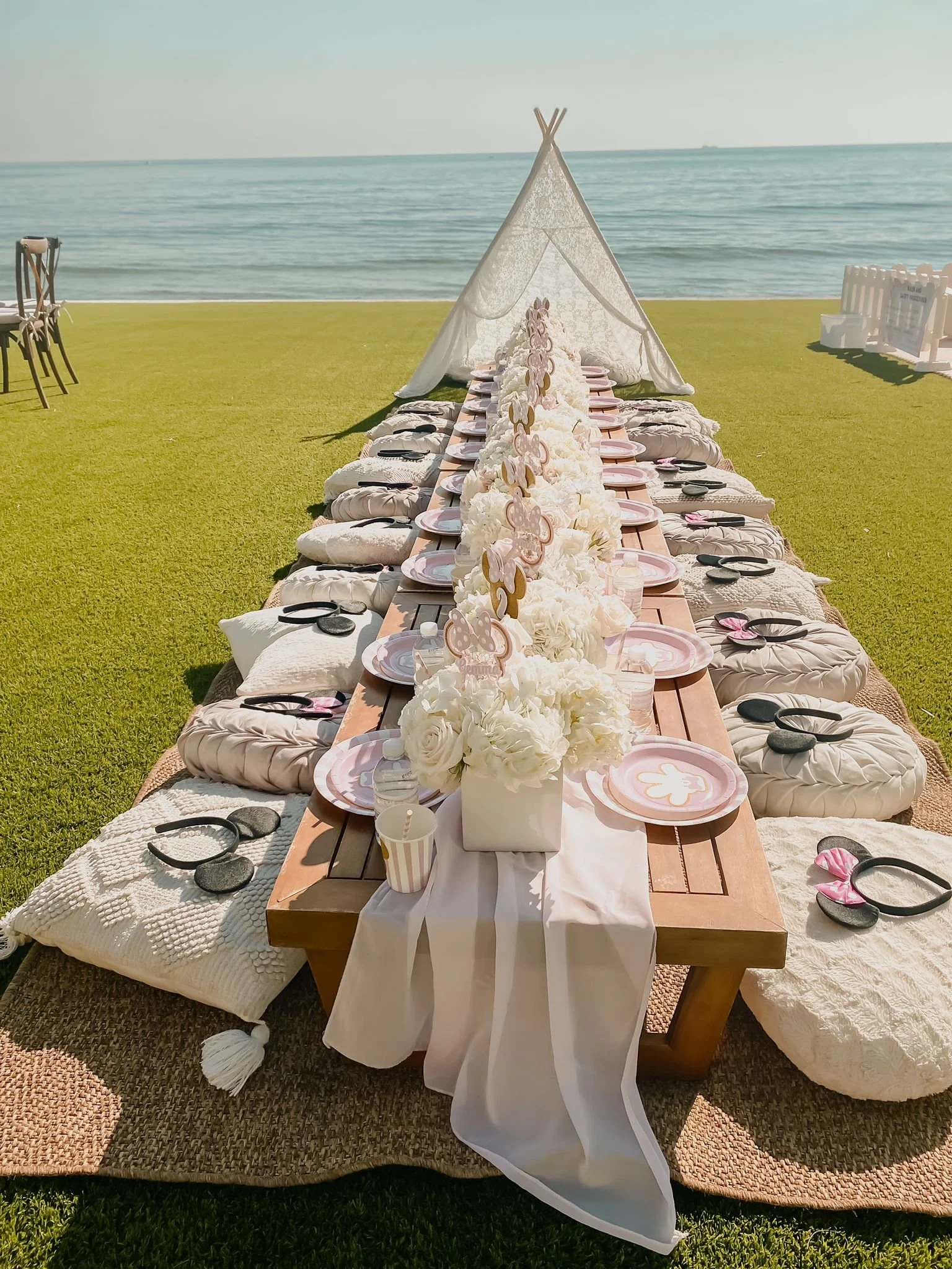 A long, low table set for a meal outdoors on a grassy area near the ocean, with a white canopy tent in the background and chairs on either side of the table.