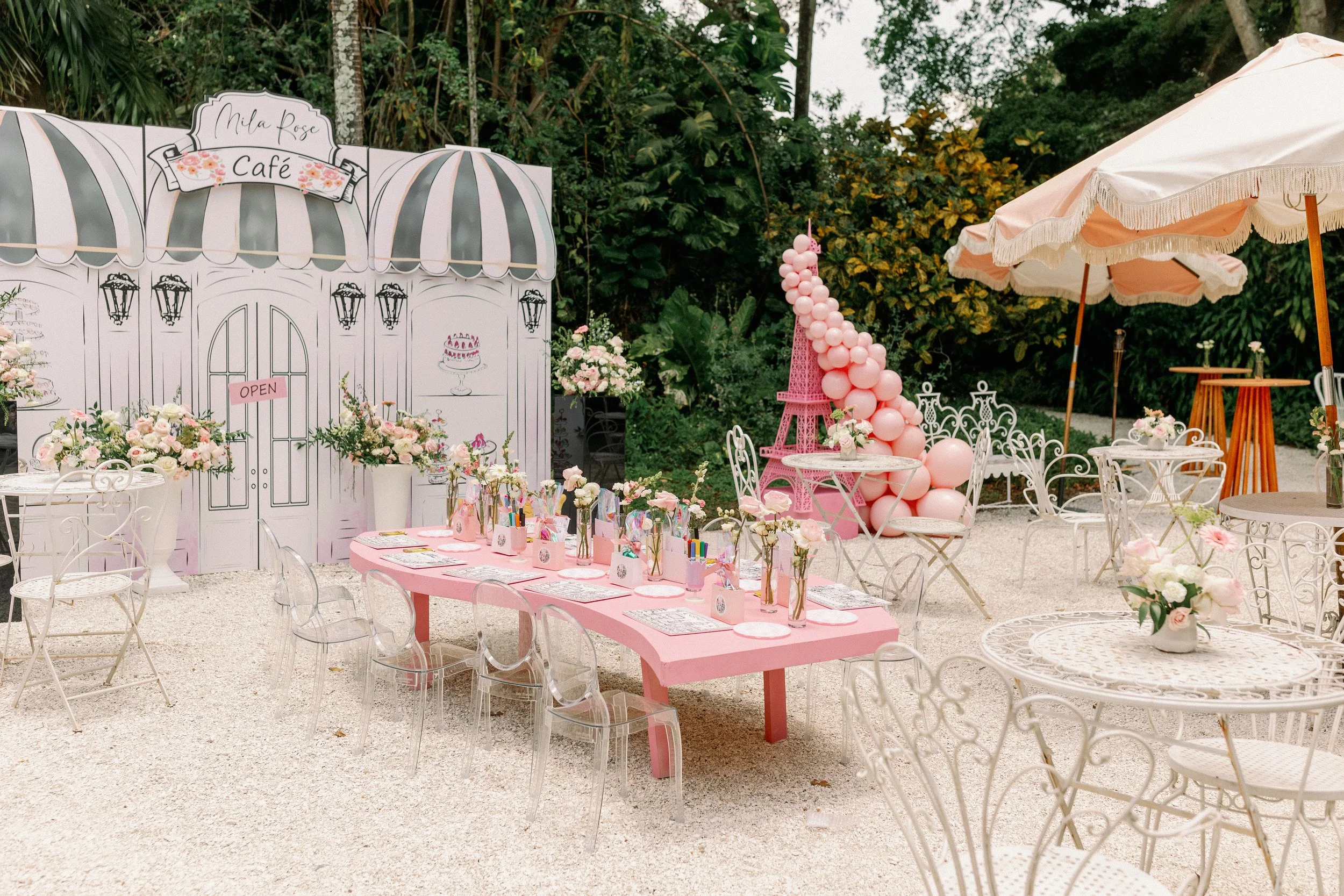 Outdoor event decorated with pink and white themes, including balloons, flowers, and a pink tabletop with party favors, set against a backdrop of greenery and white umbrellas.