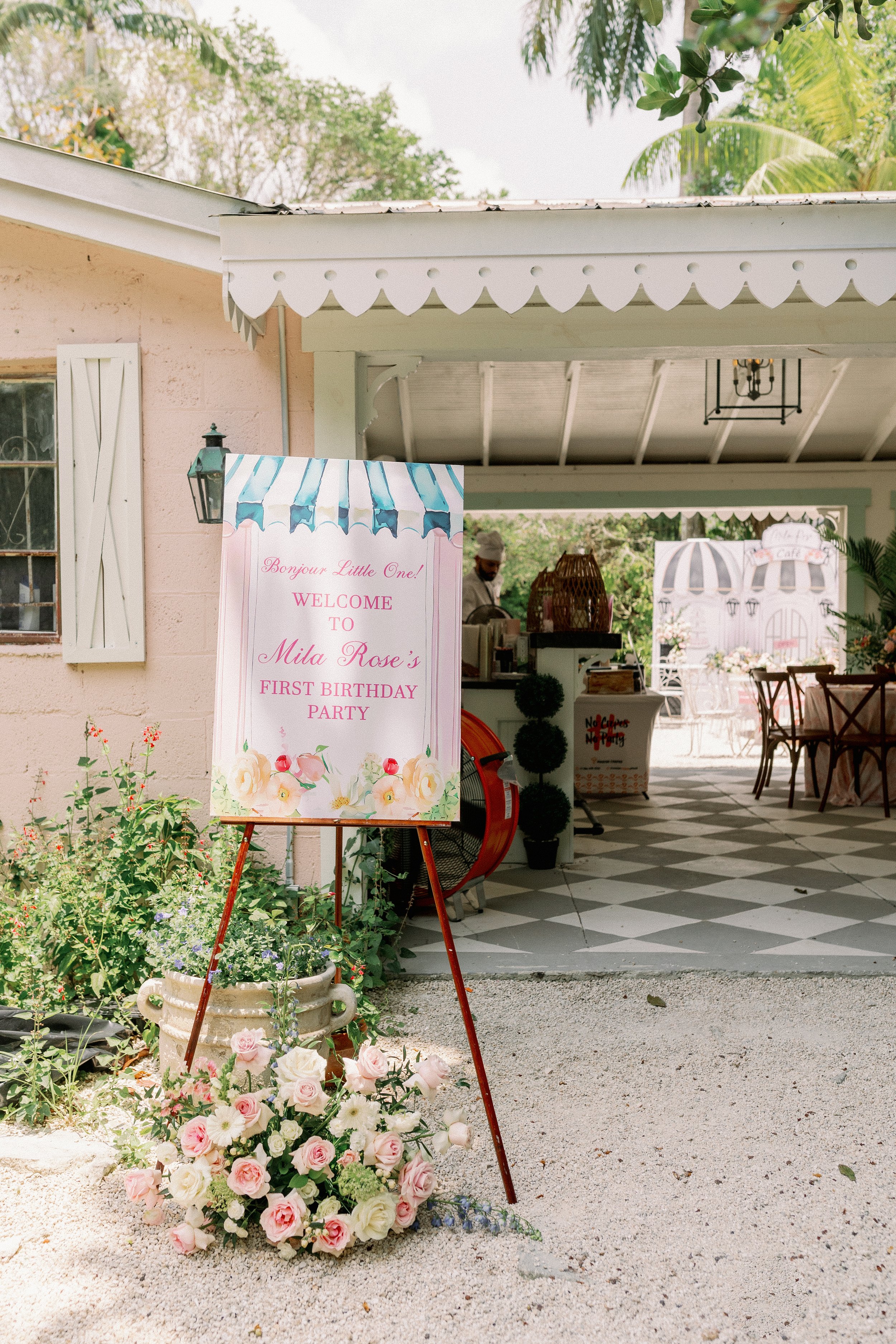 A pink sign with floral decorations and text greeting guests at a first birthday party. The sign reads: "Bonjour Little One! Welcome to Mila Rose's First Birthday Party." There is a flower arrangement with pink and white roses and some greenery in fr