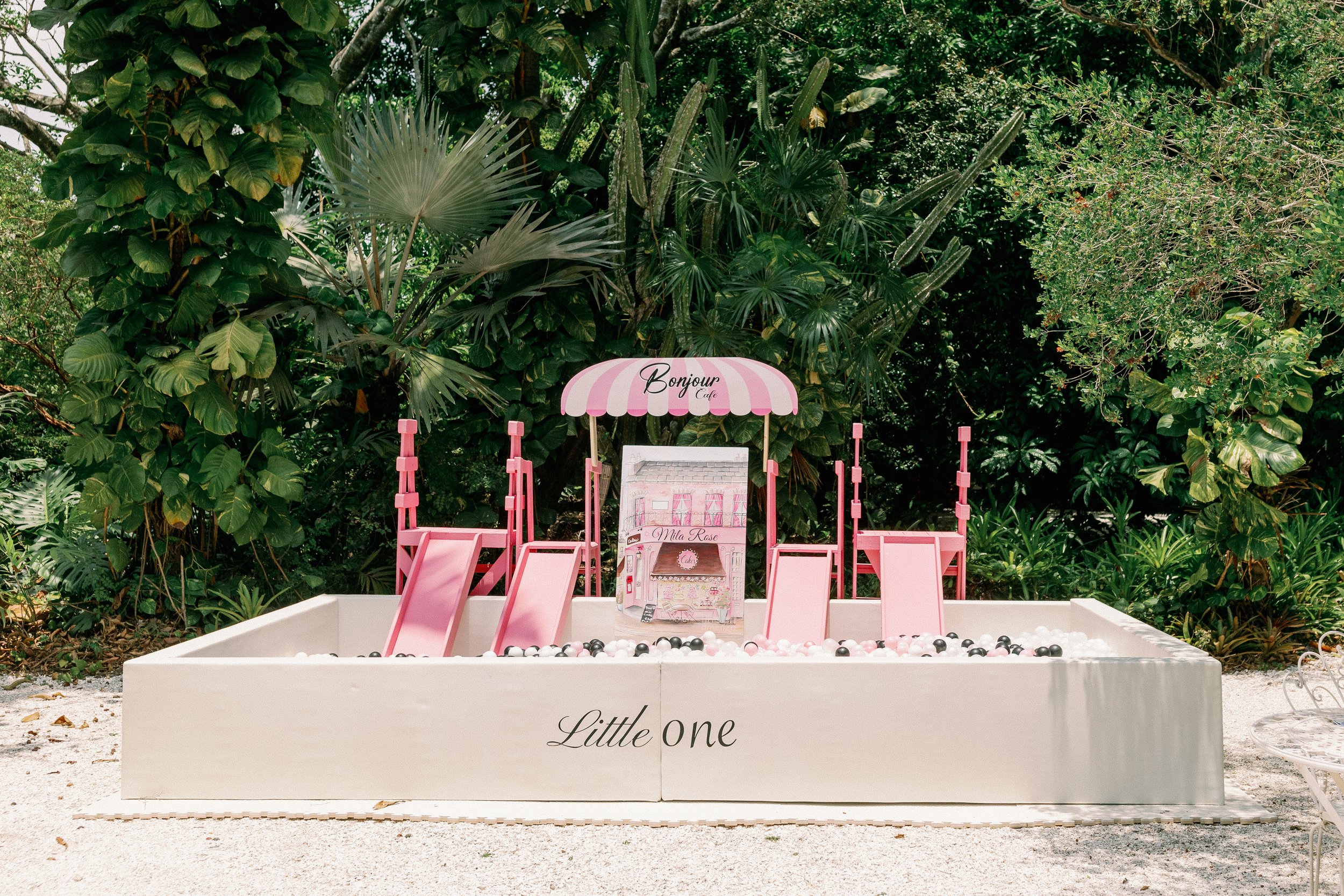 A miniature pink and white summer-themed children’s carnival stall set on a sandy outdoor area, surrounded by lush green tropical plants and trees. The stall has a striped pink and white canopy with the words "Bonjour Cafe" and a small display area b