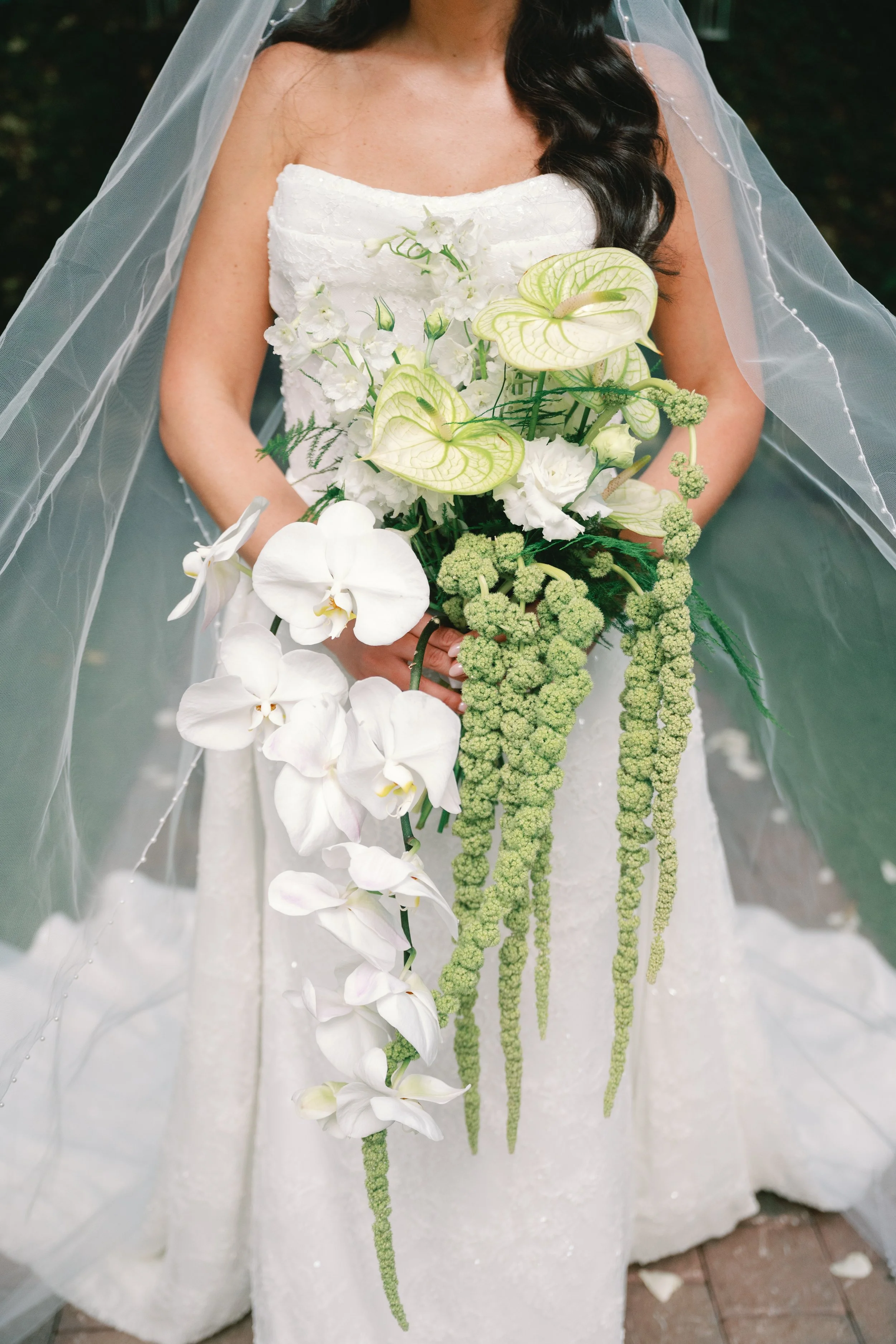 Bride holding a cascading bouquet of white orchids, anthuriums, and greenery, wearing a strapless white wedding dress and veil.