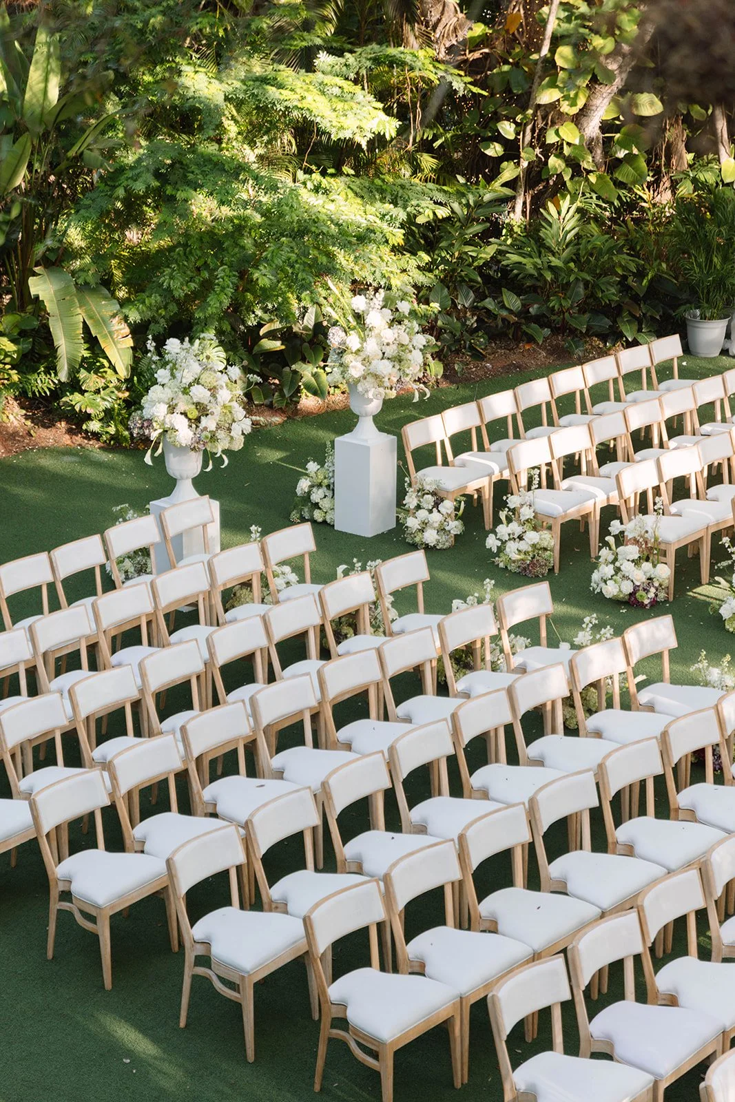 Outdoor wedding ceremony setup with white chairs, floral arrangements, and lush greenery.