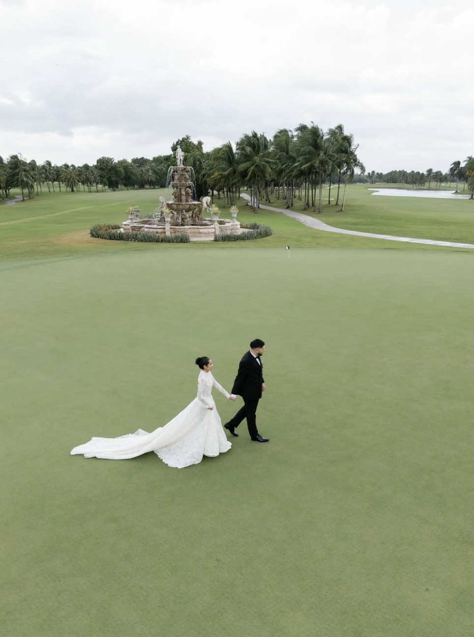 A bride and groom holding hands walking on a golf course green, with a fountain and palm trees in the background.