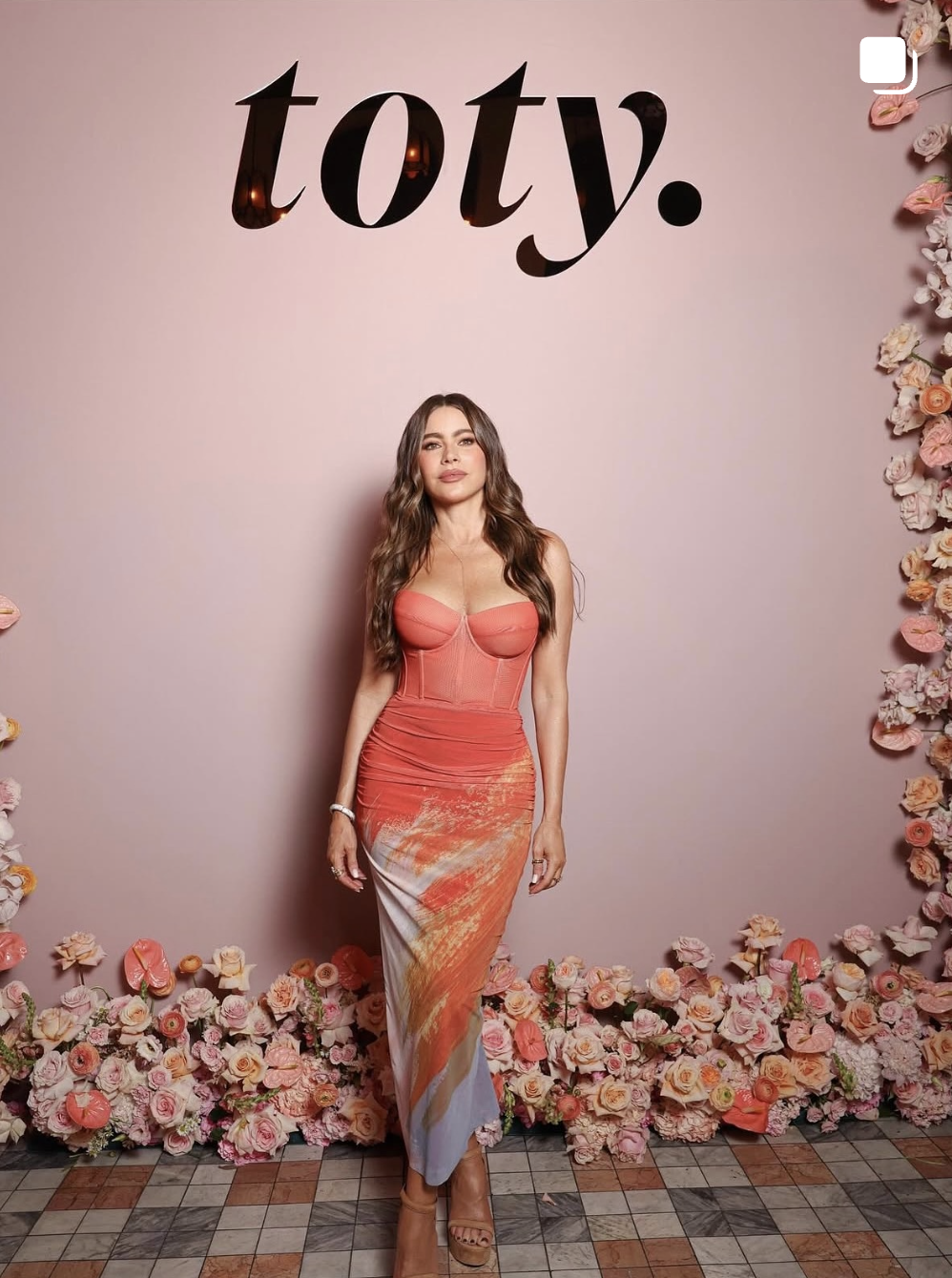 A woman in a coral and white dress standing in front of a pink wall decorated with flowers and the word 'taty' in black letters.