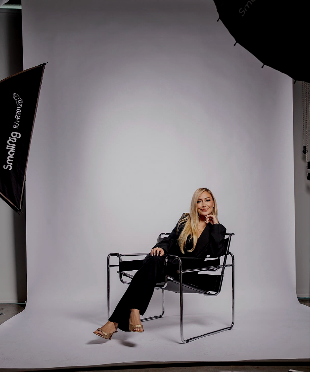 A woman with long blonde hair sitting on a black chair during a professional photo shoot in a studio with a plain grey backdrop and studio lighting equipment.