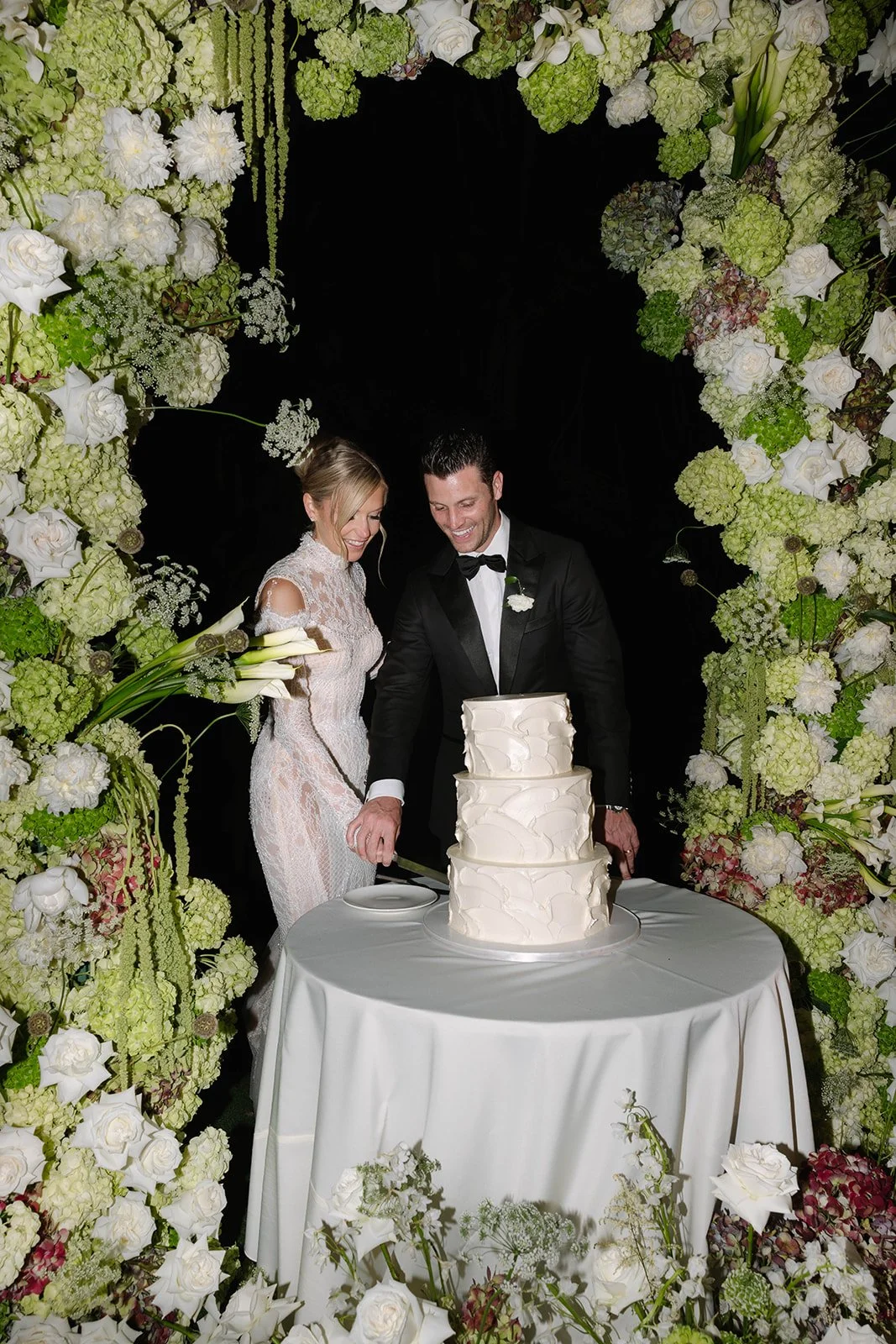 A bride and groom cut a wedding cake under a floral arch with white, green, and light purple flowers.