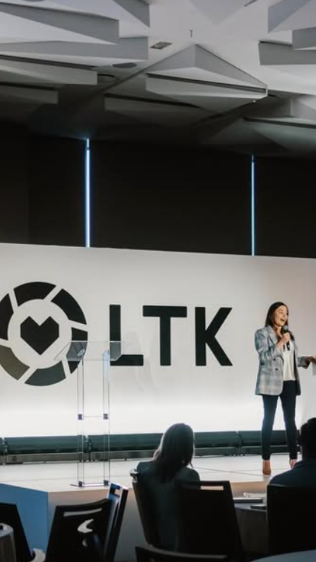 A woman speaking on stage at a conference with a large screen behind her displaying the logo 'OLTK' and a circular design.
