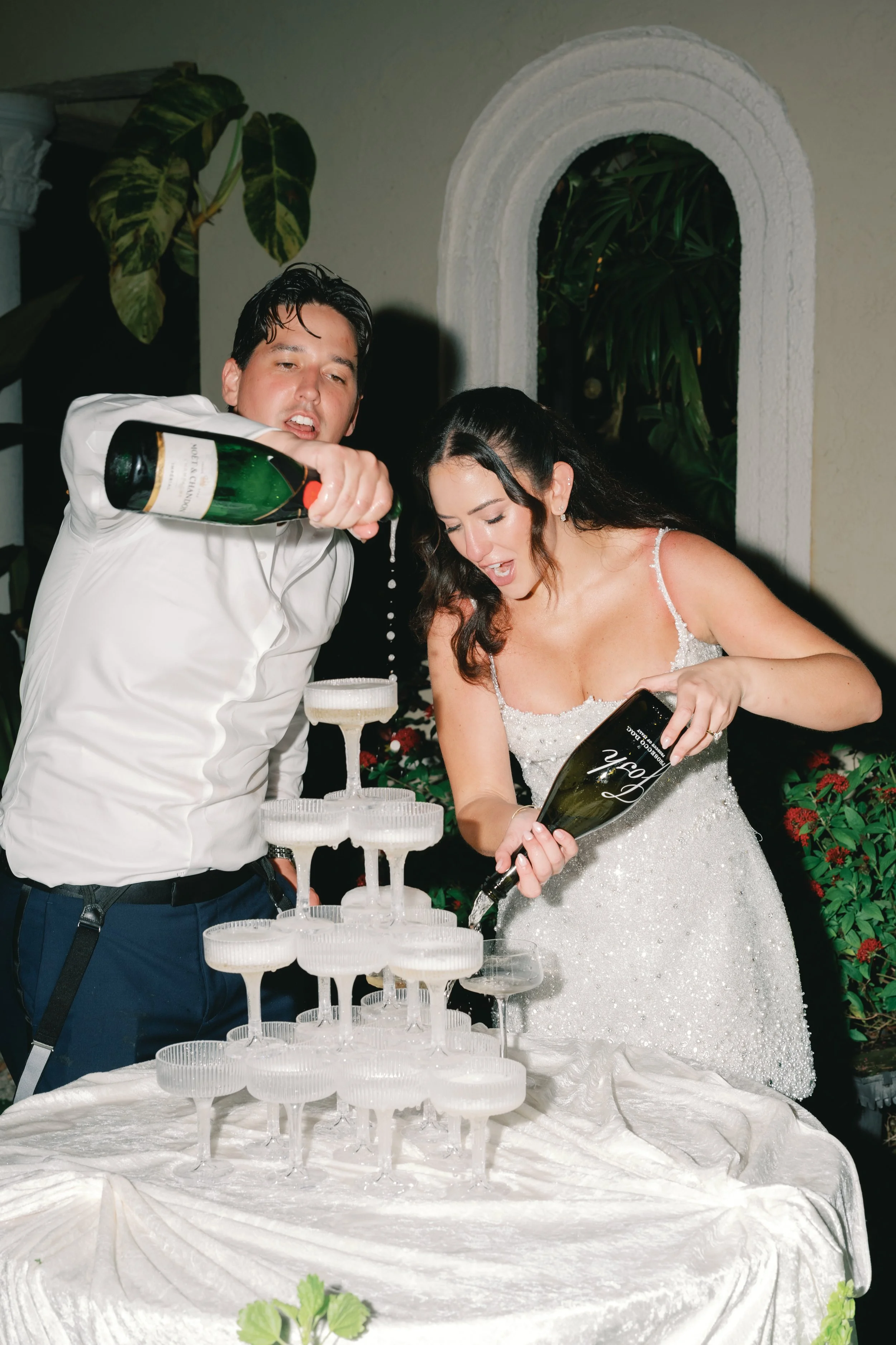 A man and woman in formal attire pouring champagne into a multi-tiered champagne tower at a celebration event.