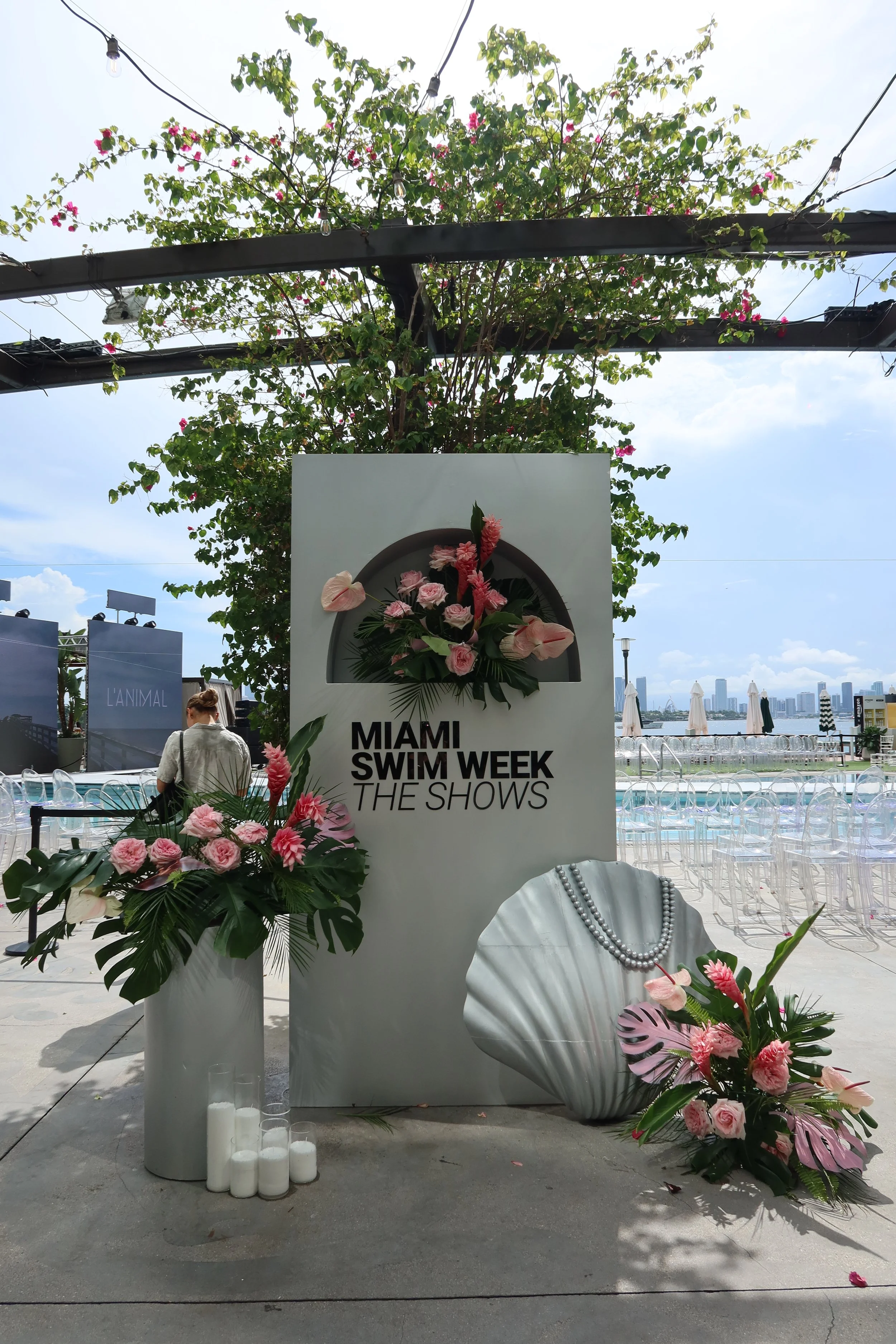 Decorative display for Miami Swim Week featuring a large white sign with floral arrangements of pink and white flowers, green tropical leaves, and a pearl necklace on a stylized figure, set outdoors near a water view with city skyline in the backgrou