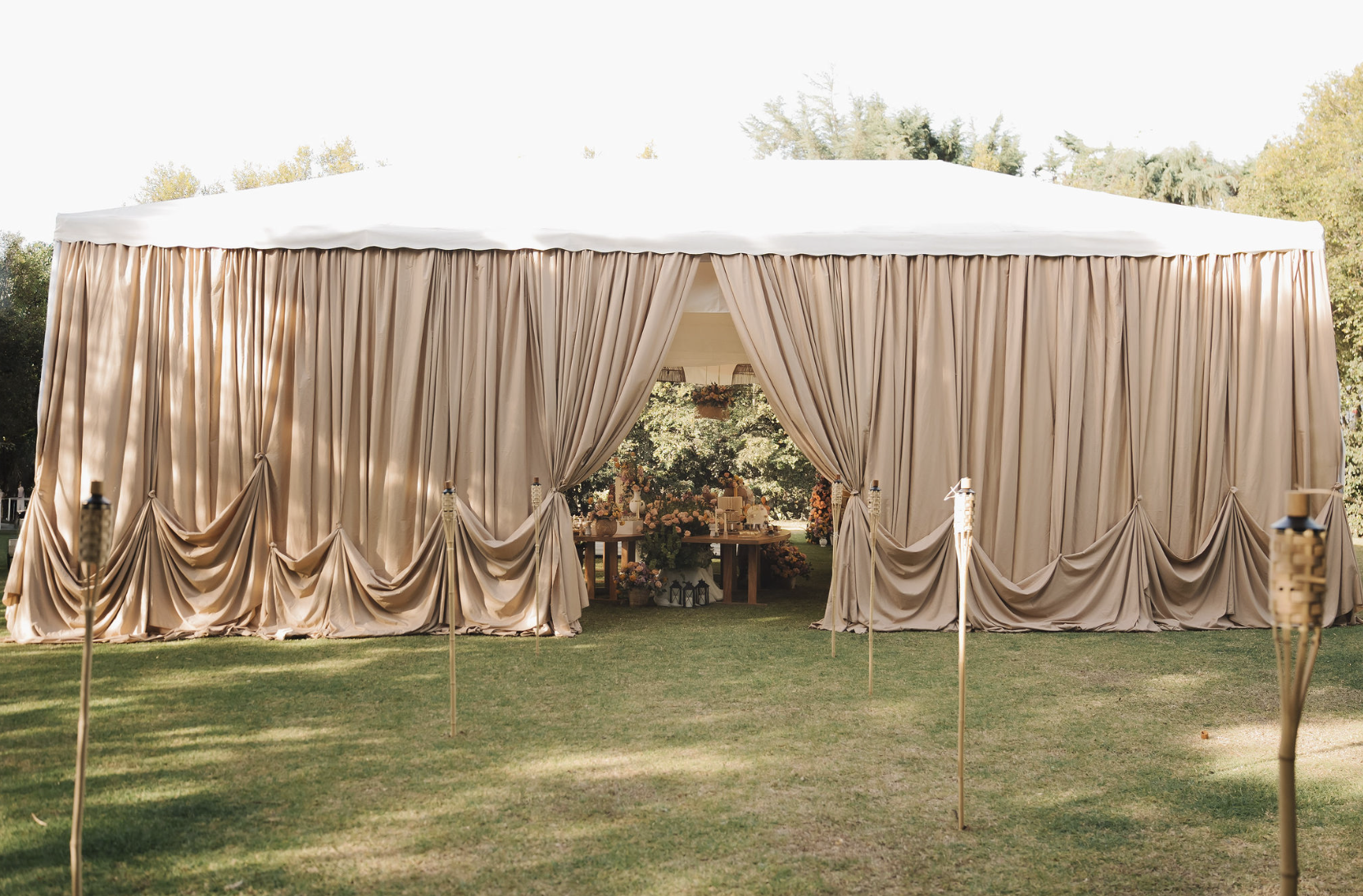A large beige tent with closed curtains on the sides, set up outdoors on green grass, with trees in the background, decorated for an event or celebration.