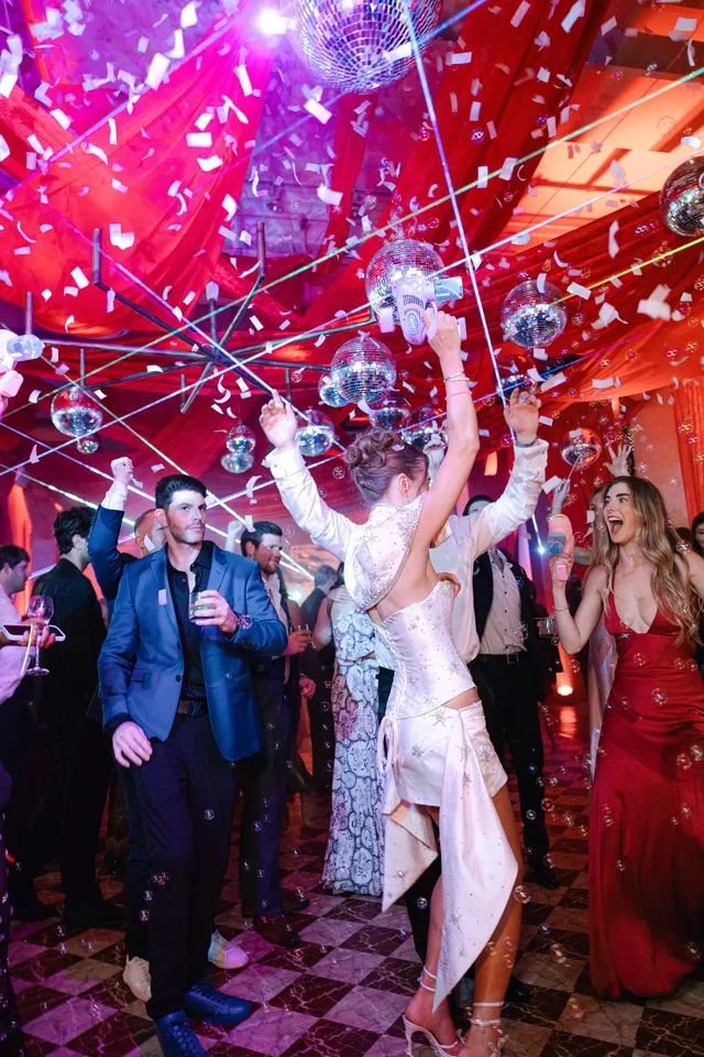 People dancing and celebrating at a party with disco balls, confetti, and colorful lighting.