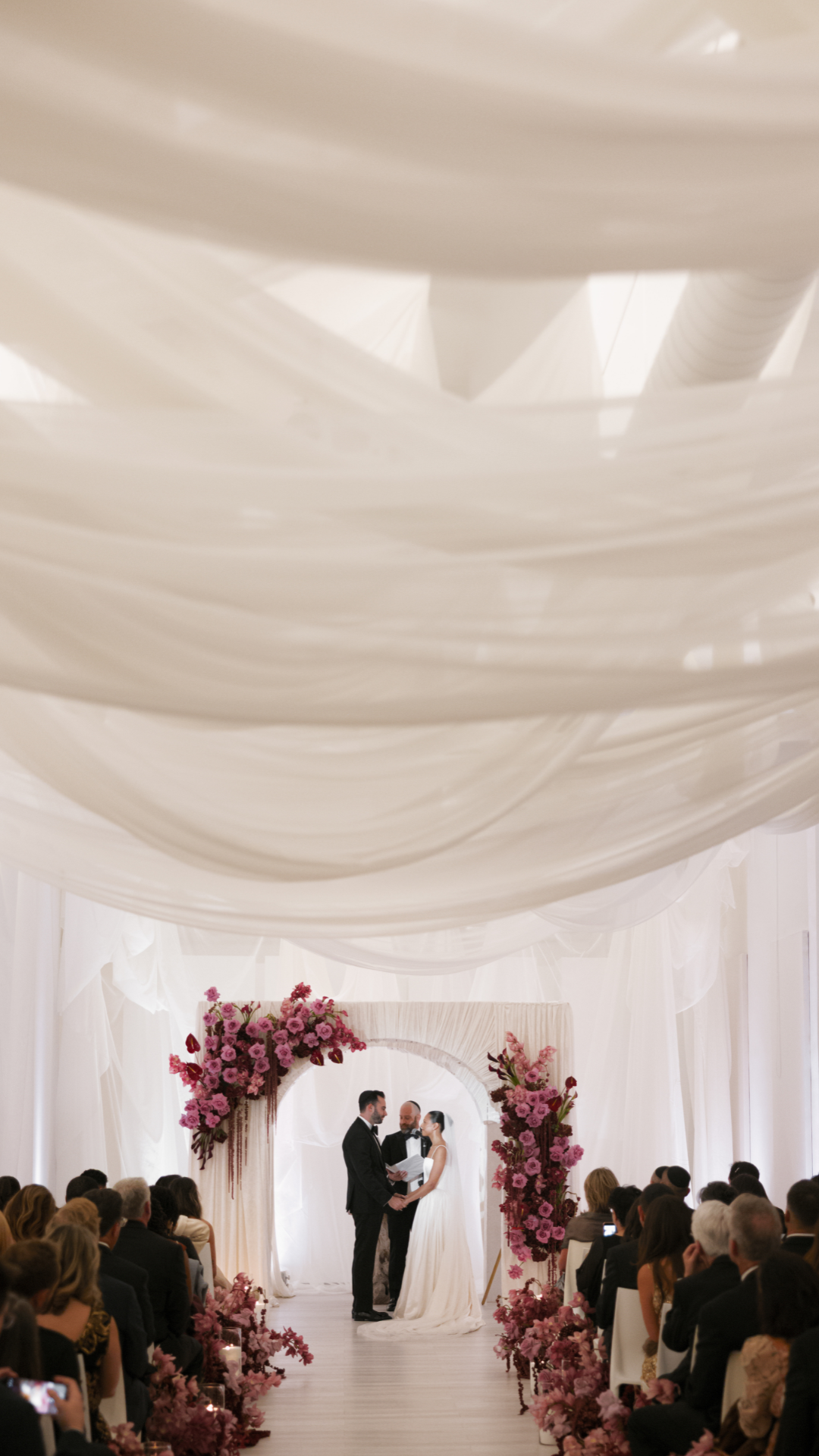 A wedding ceremony with a bride and groom exchanging vows in front of a floral arch, surrounded by guests in an elegant, draped venue with pink and purple flowers.