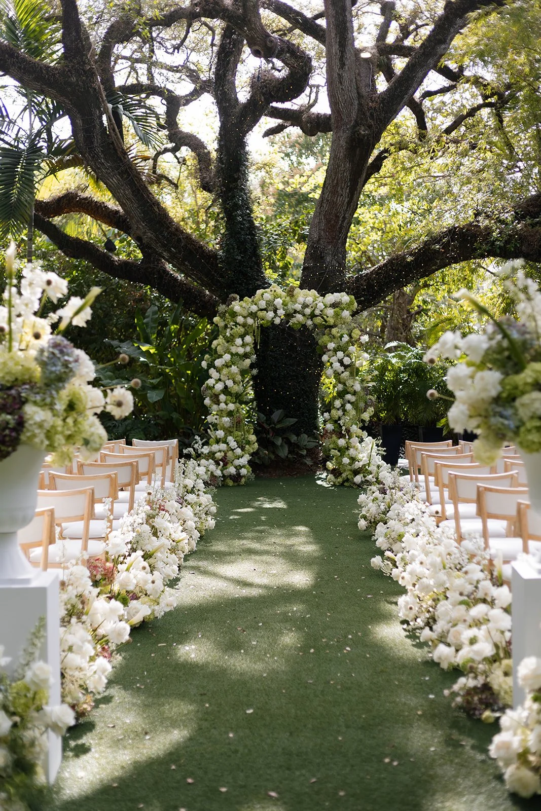 Outdoor wedding ceremony setup with white chairs and floral decorations under a large tree with an arch made of white flowers at the end of the aisle.