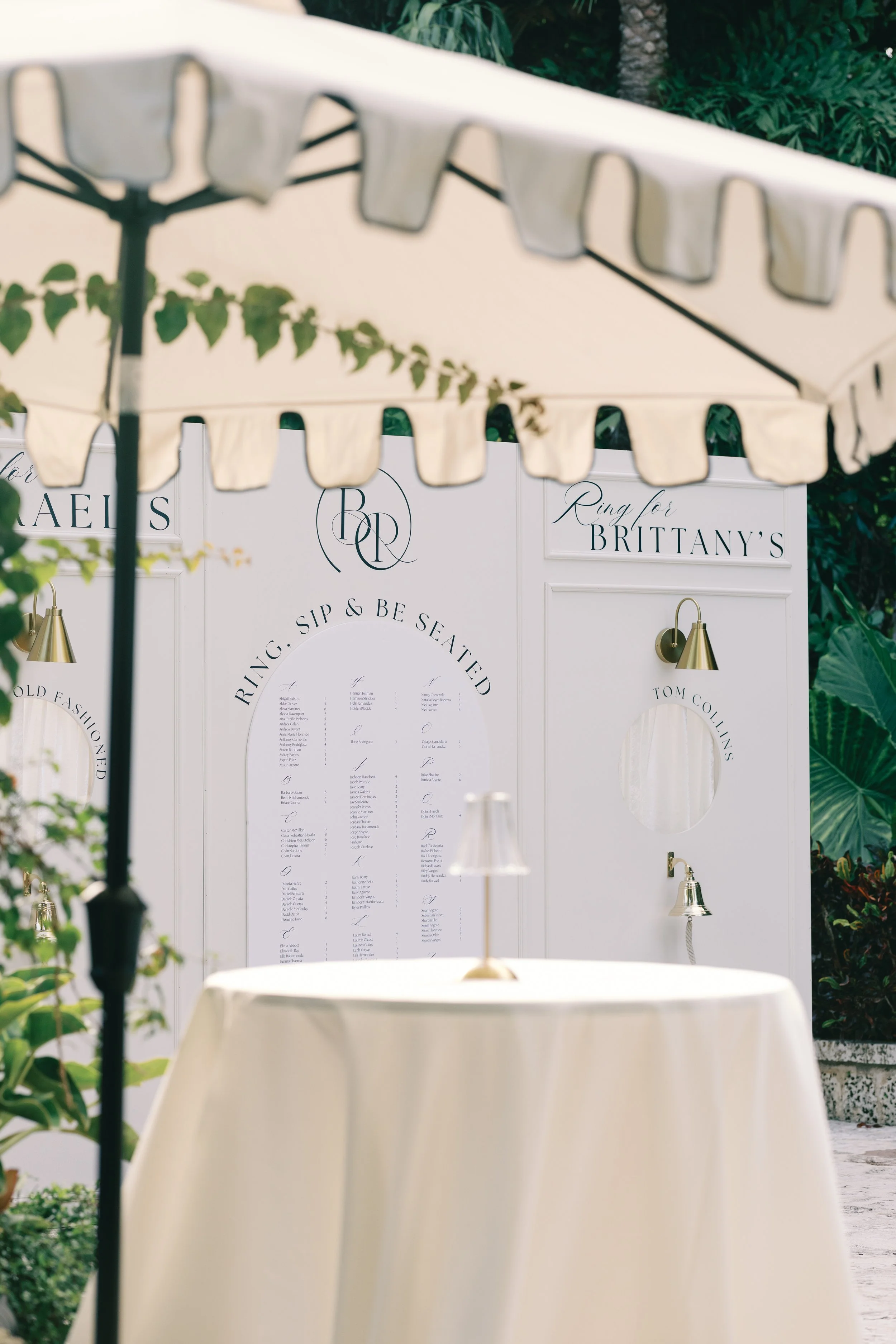 A decorative outdoor display with a white signboard that has elegant black text and gold lamps, surrounded by greenery, with umbrellas in the foreground.